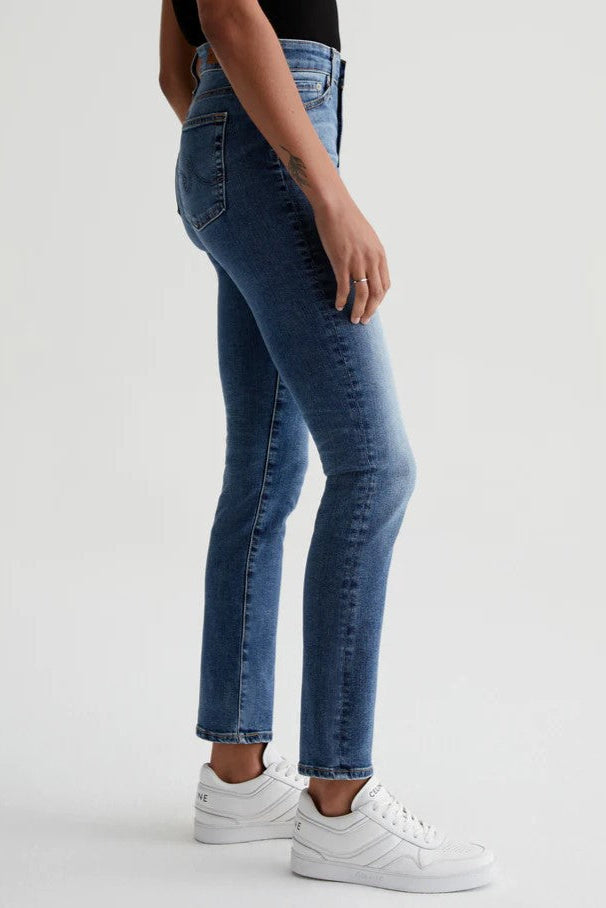 A person in AG Jeans’ Mari Straight Jean in 15 Years Shoreline, a black top, and white sneakers stands sideways against a plain light background with one arm relaxed by their side.