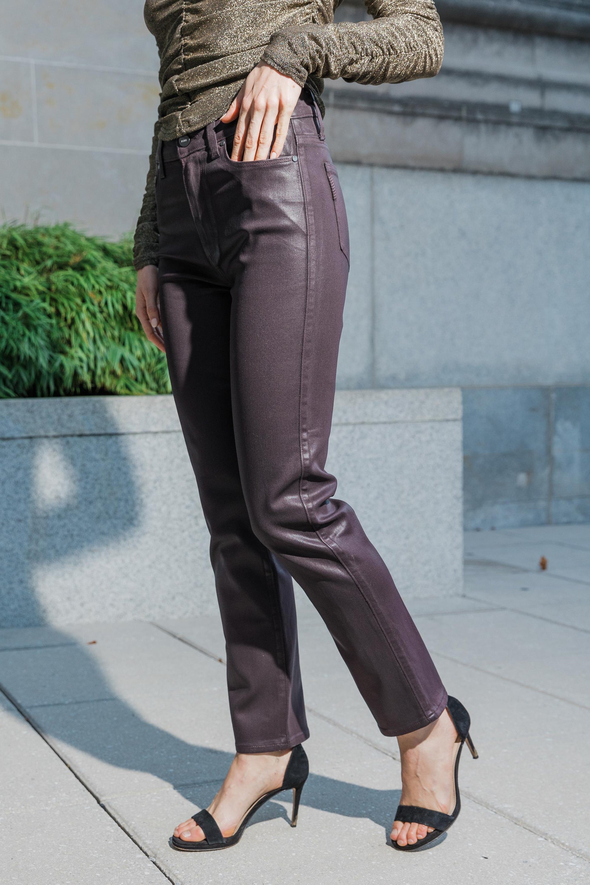 A person wears the Paige Cindy Jean in Black Cherry, paired with a shimmery green long-sleeve top and black heeled sandals. They stand outdoors on a paved area near a stone wall and green shrubbery, with their face not visible.