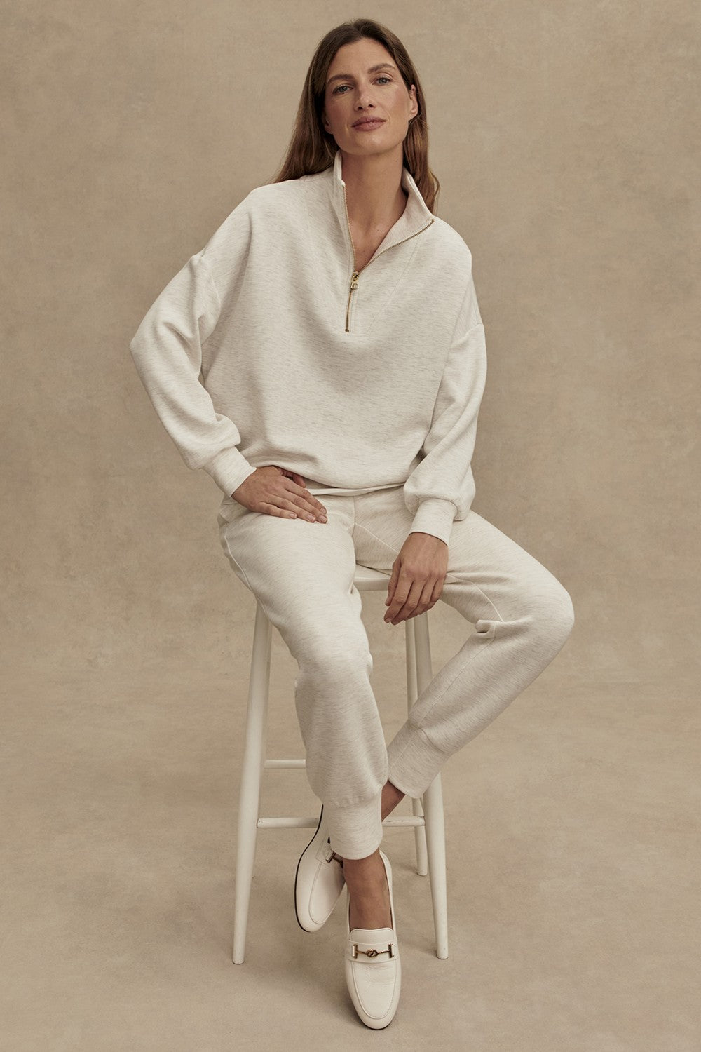 A woman wearing the Varley Hawley Half Zip Sweatshirt in Ivory, crafted from plush DoubleSoft fabric, paired with matching joggers and white loafers, sits on a white stool against a beige studio backdrop, looking calmly at the camera.