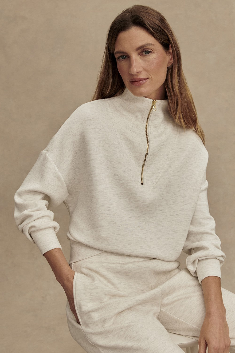 A woman with long brown hair wears the Varley Hawley Half Zip Sweatshirt in Ivory Marl, paired with matching pants, standing with one hand in her pocket against a neutral background.