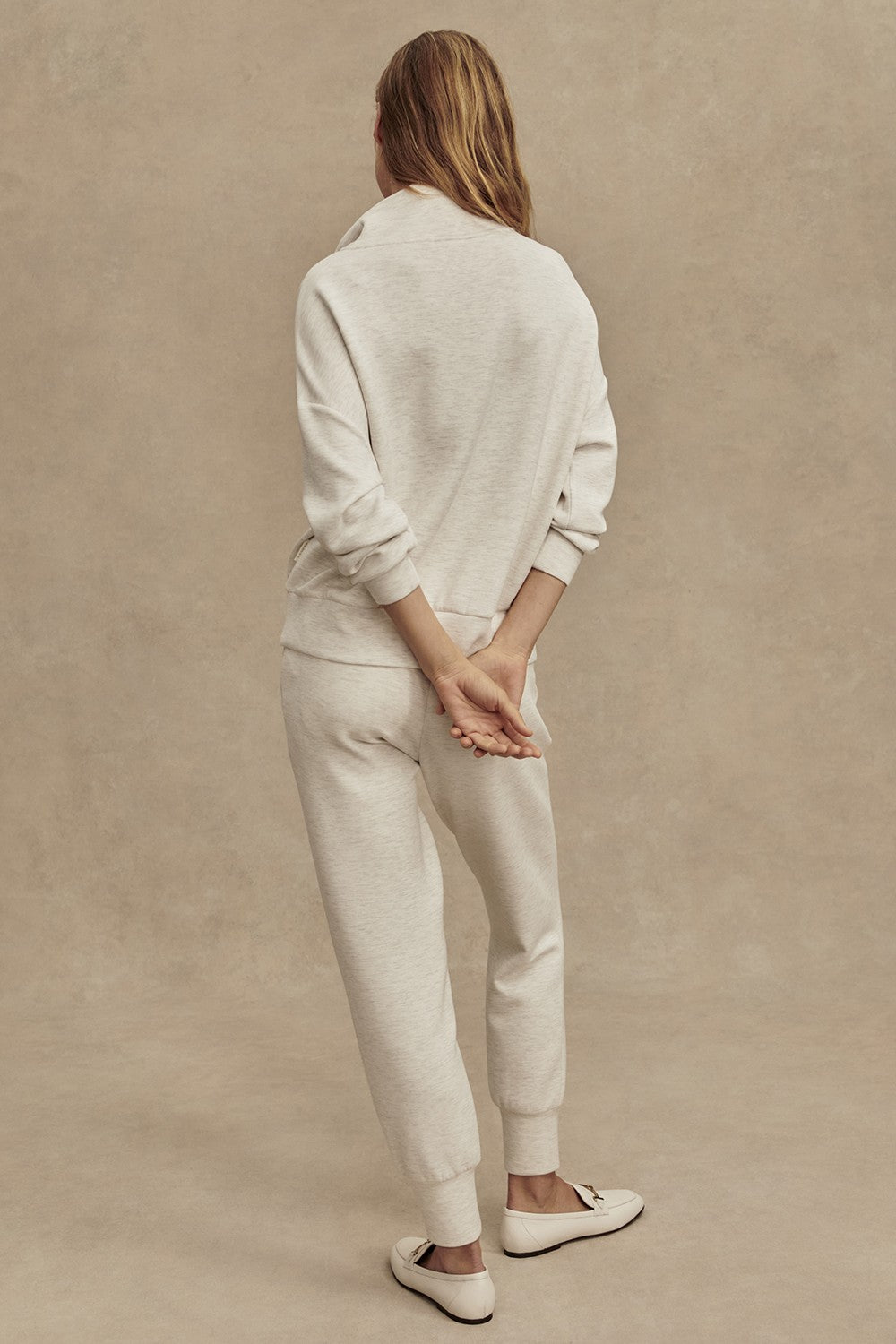 A person with long hair, dressed in the Varley Hawley Half Zip Sweatshirt in Ivory—crafted from soft DoubleSoft fabric—and matching sweatpants, stands barefoot with their hands clasped behind their back, facing away from the camera against a plain beige background.