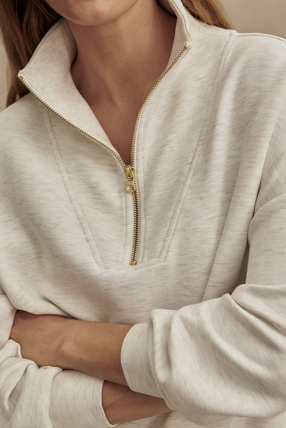A person wears the Varley Hawley Half Zip Sweatshirt in Ivory, featuring a gold zipper and crafted from soft DoubleSoft fabric. With arms folded, the upper body is in focus to highlight the textured material and zipper detail, while the person's face is partially out of frame.