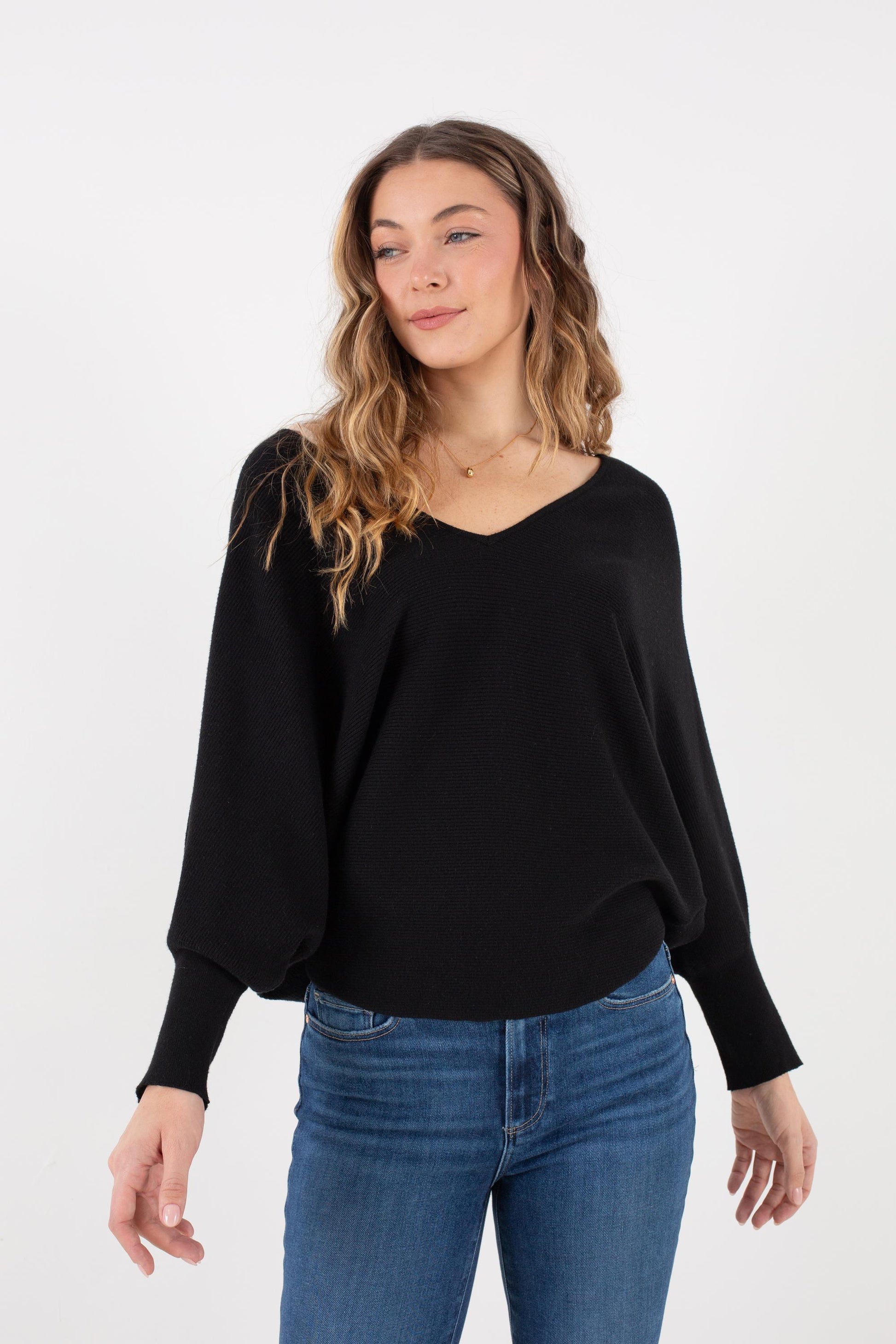 A woman with wavy hair wears the Kerisma Dolman Ryu V Neck in Black, paired with blue jeans, standing against a plain white background. She looks off to the side with a slight smile.
