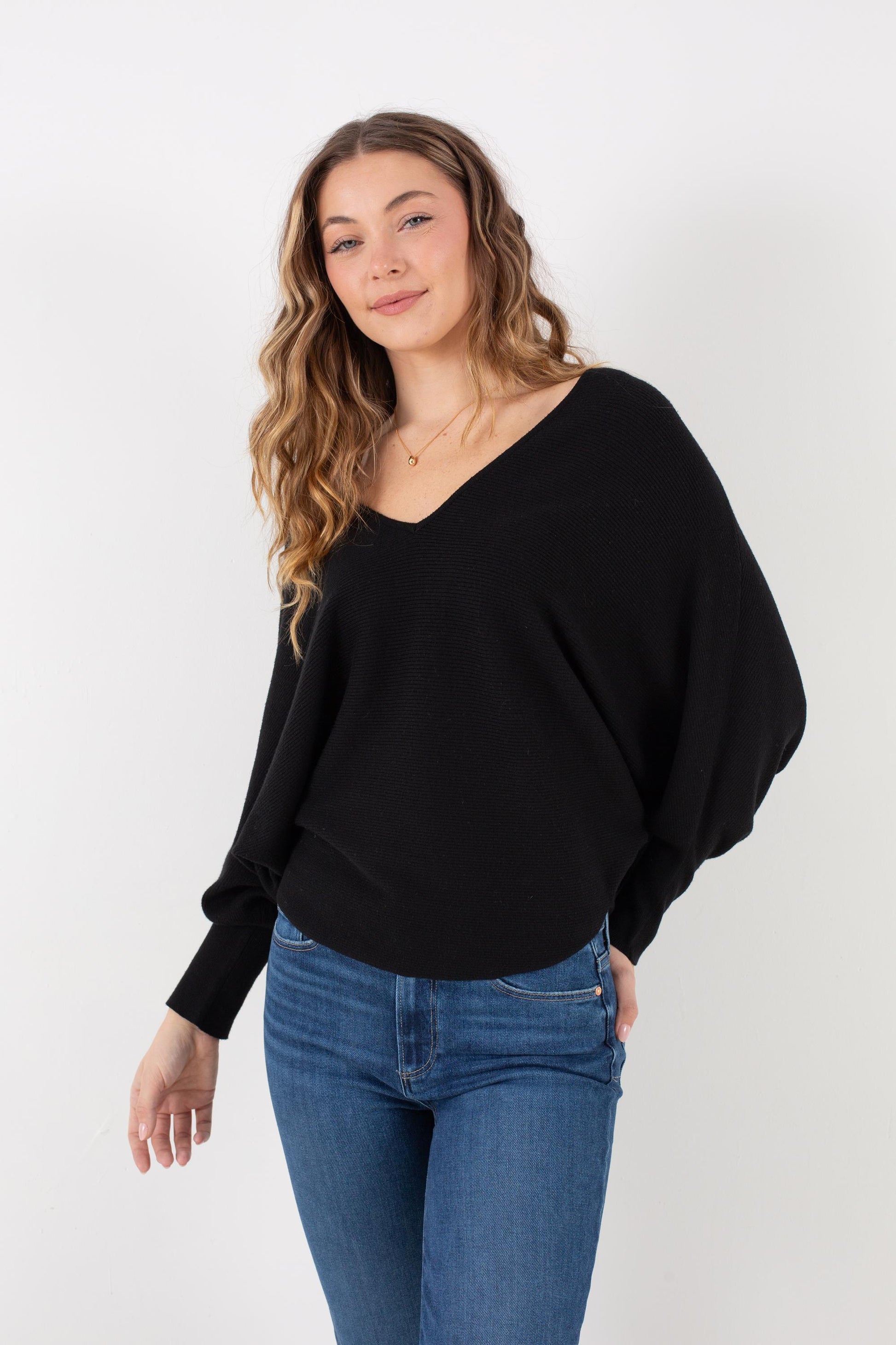 A woman with wavy brown hair stands against a plain white background, smiling slightly as she wears the Kerisma Dolman Ryu V Neck in Black paired with blue jeans.