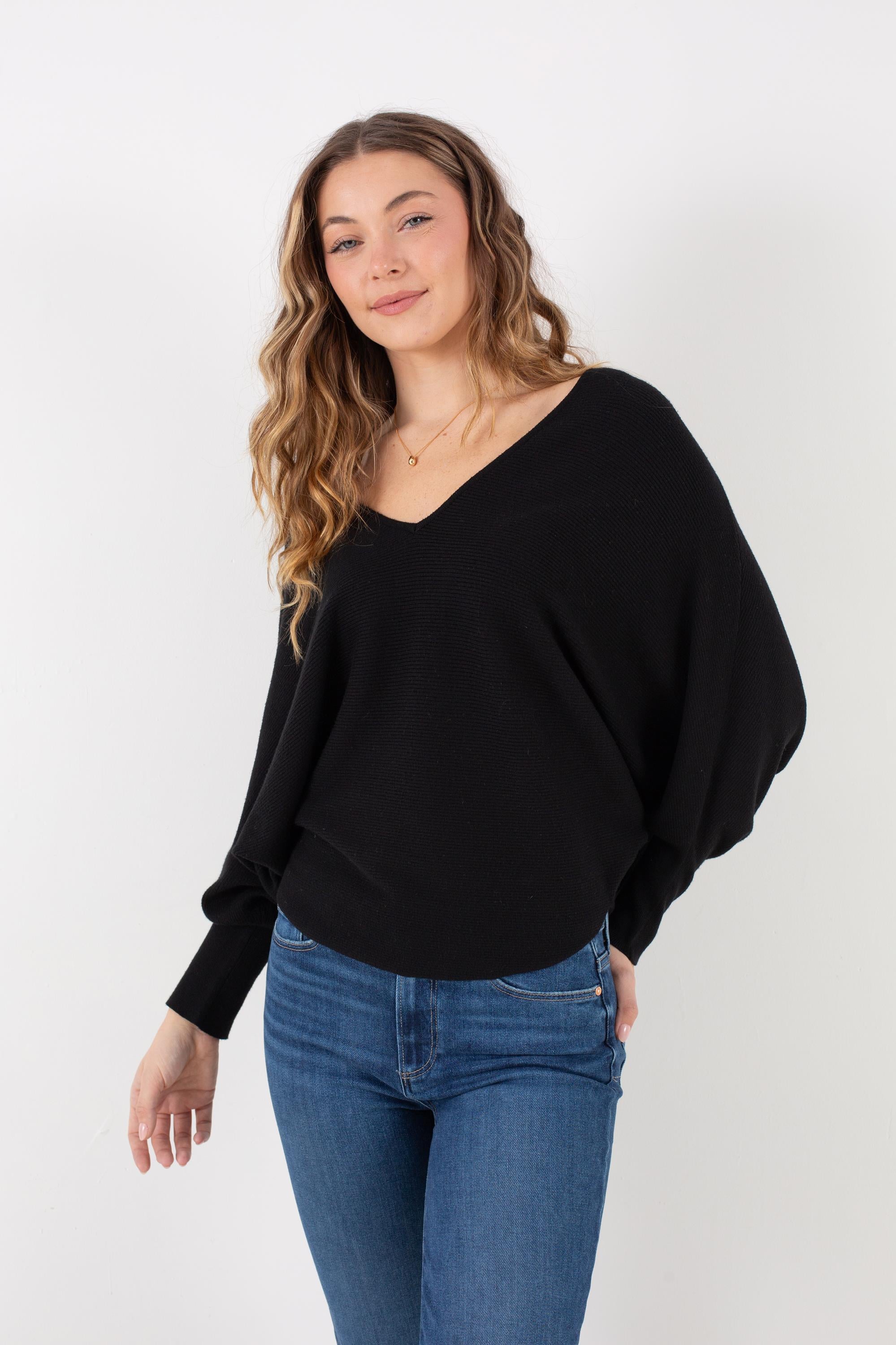 A woman with wavy brown hair stands against a plain white background, smiling slightly as she wears the Kerisma Dolman Ryu V Neck in Black paired with blue jeans.