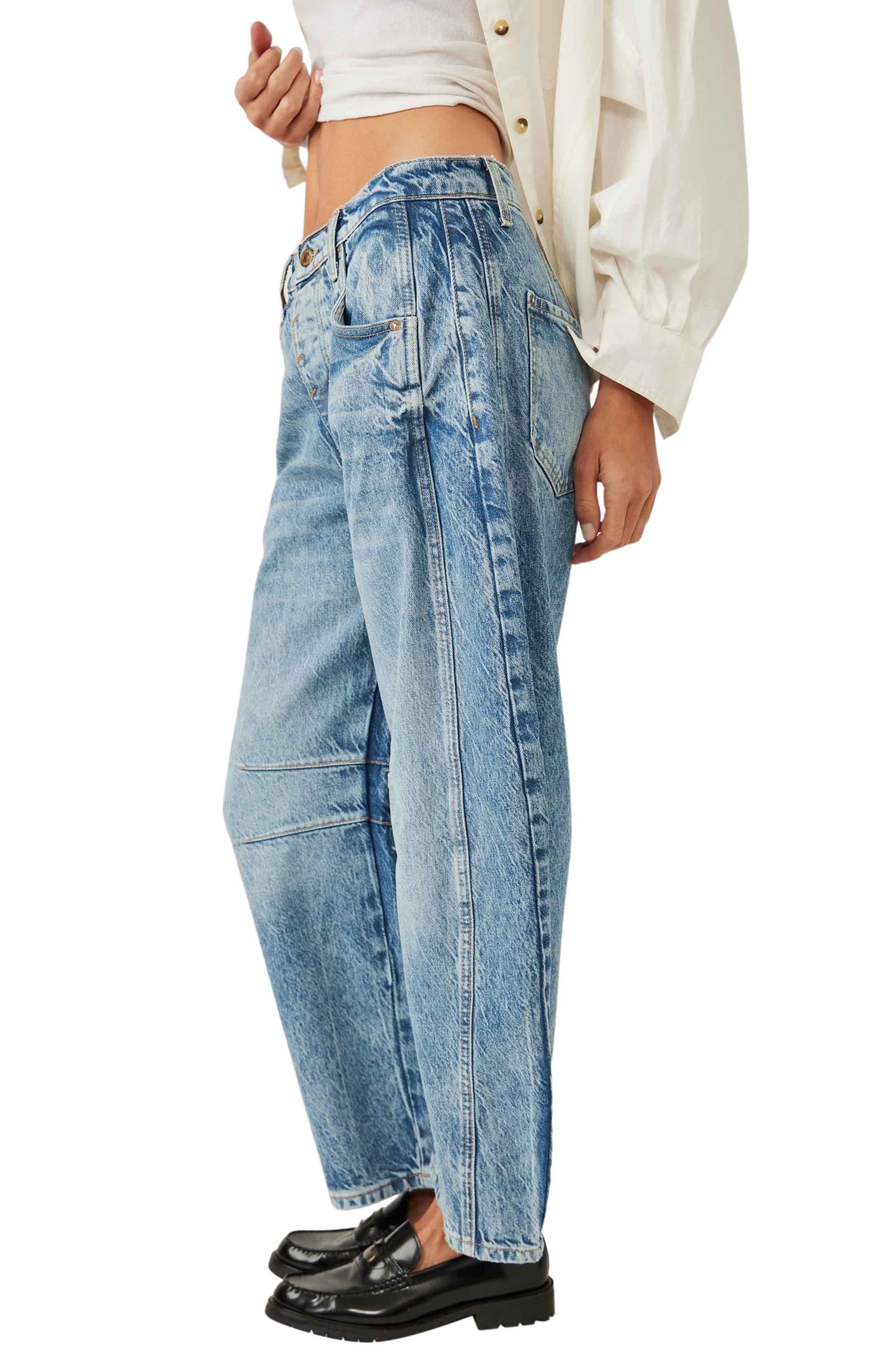 A person wearing the Good Luck Mid Rise Barrel jeans by Free People—light blue, high-waisted, relaxed-fit cotton with five pockets and visible seams—paired with a white shirt and black loafers, shown from the side against a white background.