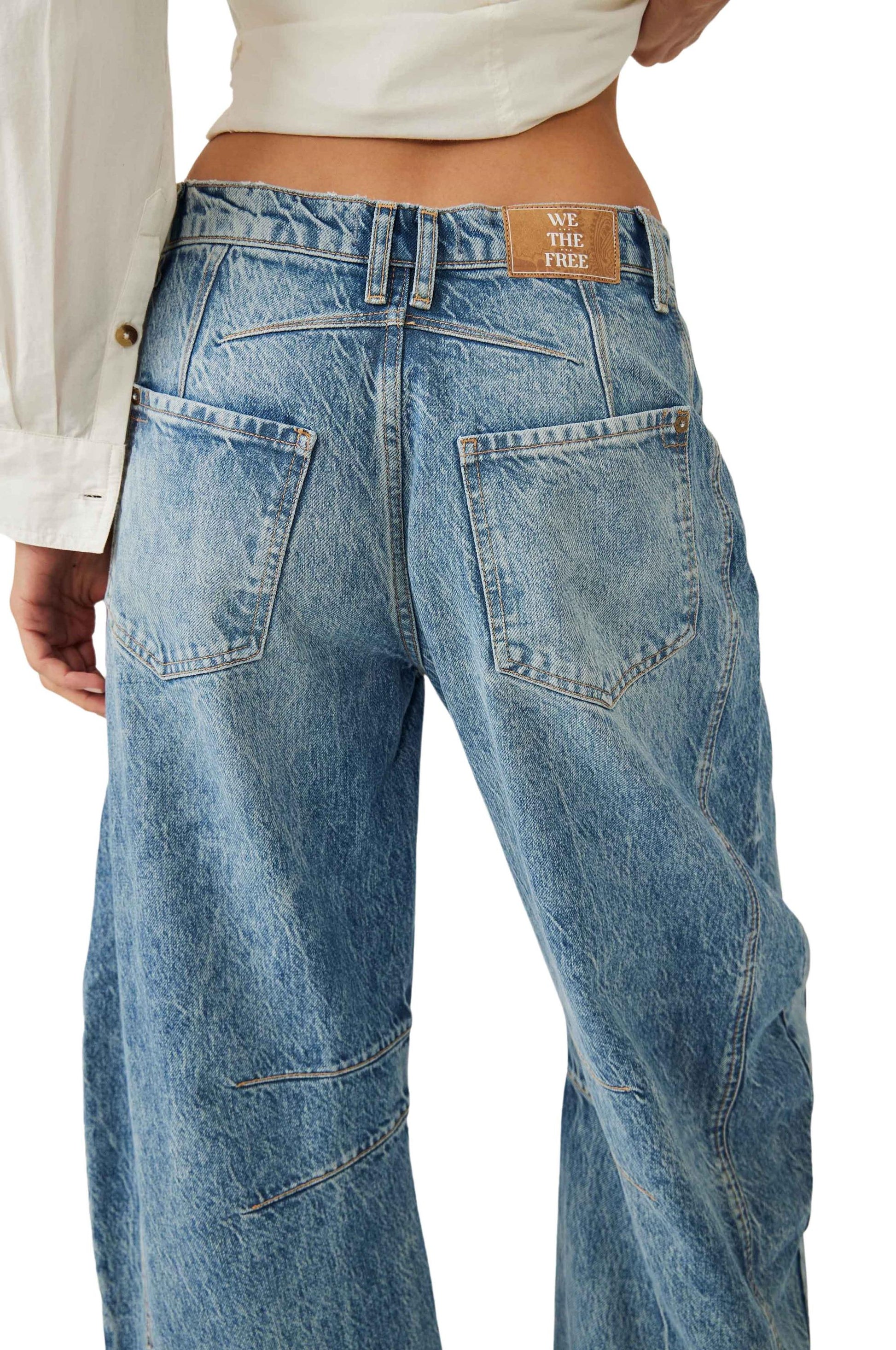 A person wears Free People Good Luck Mid Rise Barrel jeans in light blue—wide-leg, five-pocket style with visible seams and a brown We The Free label—paired with a white long-sleeve shirt. These 100% cotton jeans are made in Turkey.