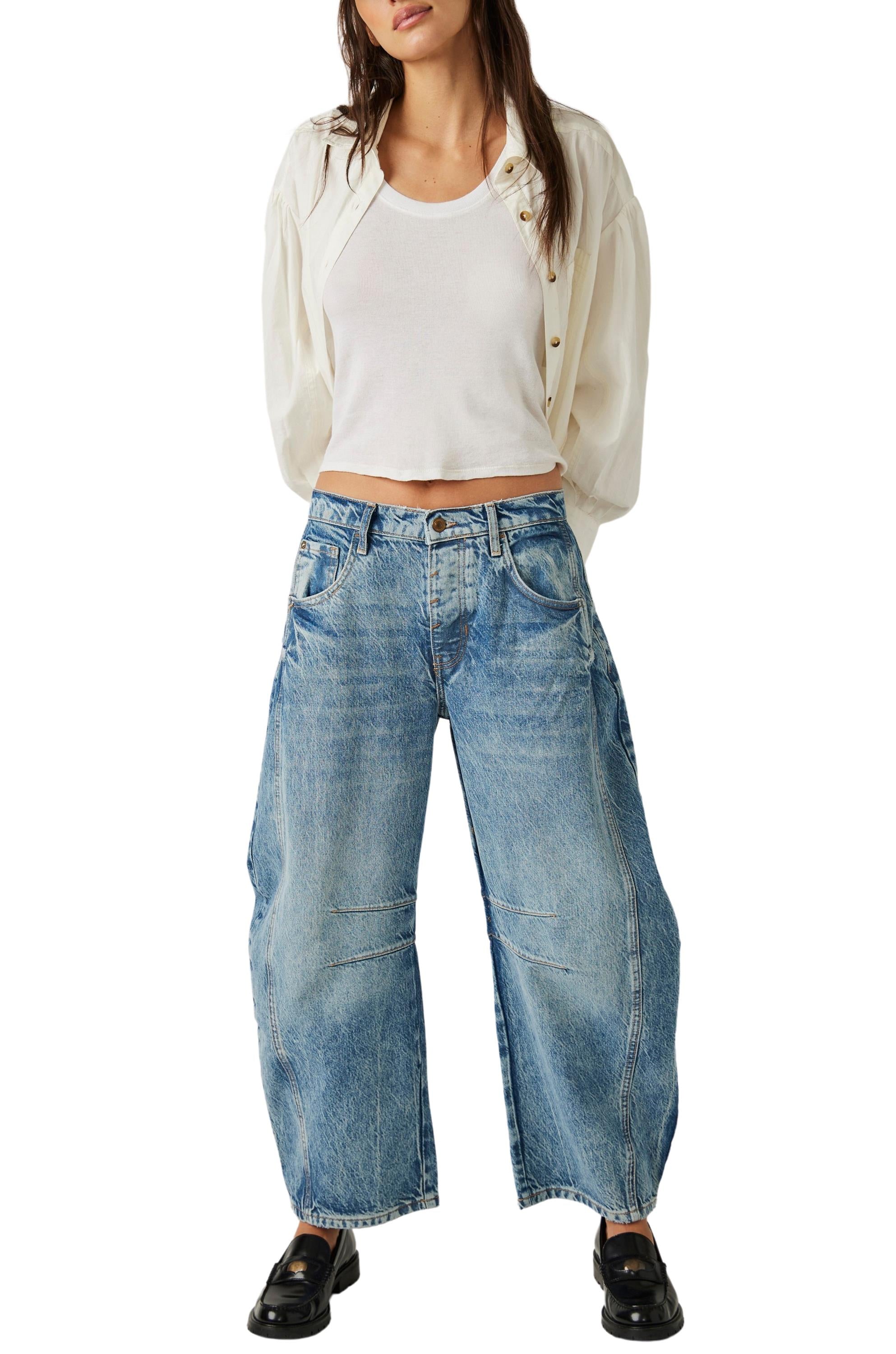 A person in a loose white shirt over a tank top, black shoes, and Free People Good Luck Mid Rise Barrel wide-leg jeans in light denim stands with hands behind their back against a white background.