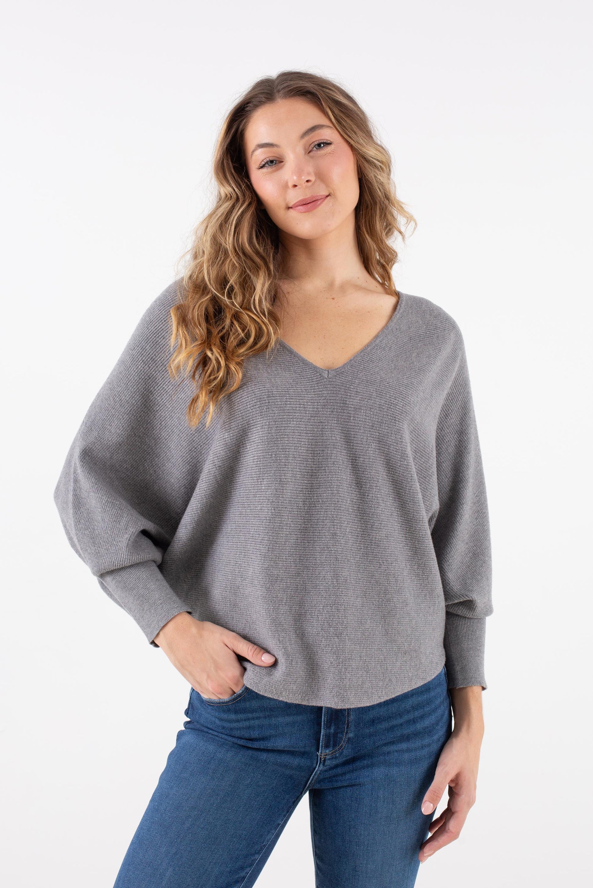 A woman with long wavy hair wears the Kerisma Dolman Ryu V Neck in Ash Grey, paired with blue jeans, standing against a white background with one hand in her pocket and a slight smile.