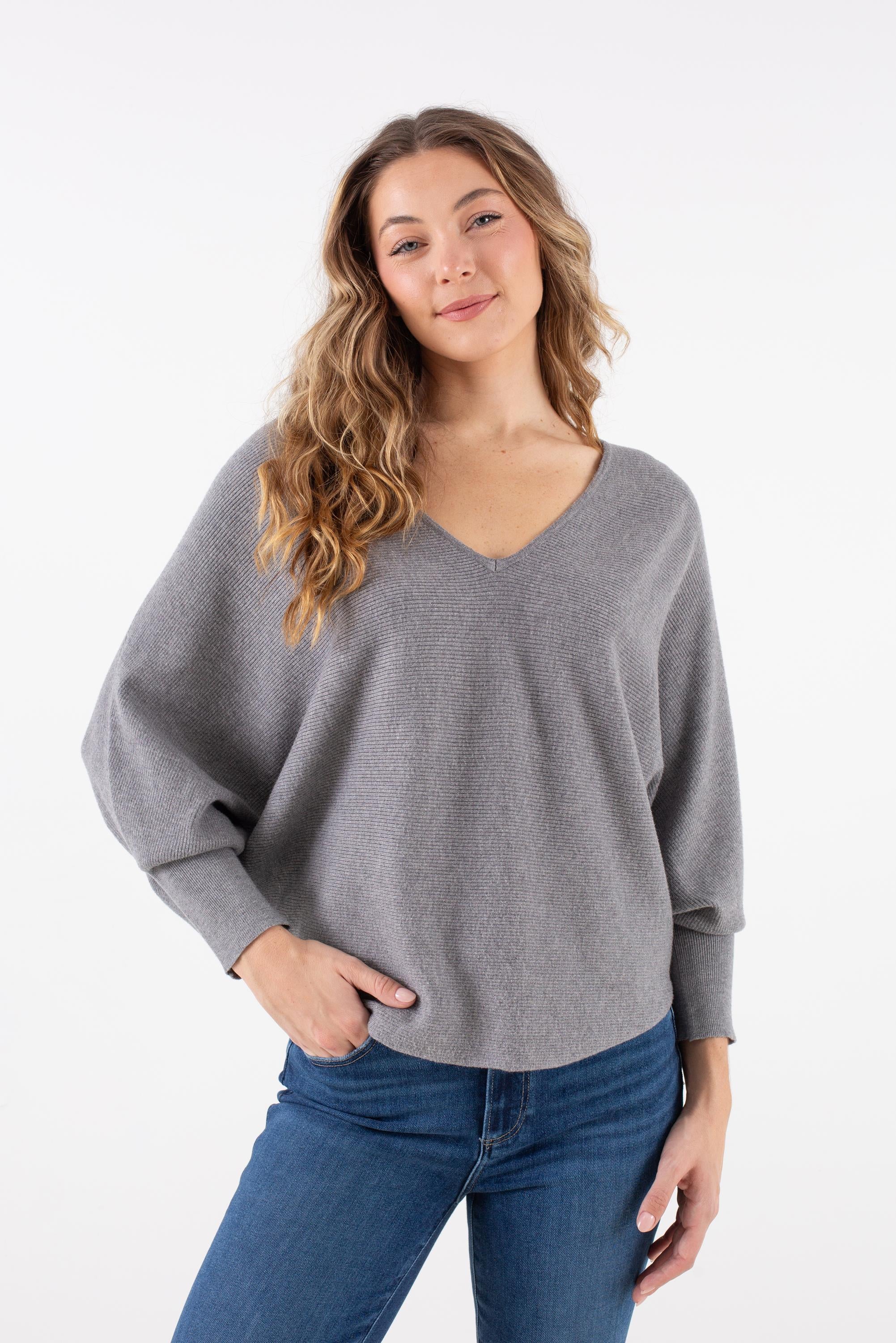A woman with long wavy hair wears the Kerisma Dolman Ryu V Neck in Ash Grey, paired with blue jeans, standing against a white background with one hand in her pocket and a slight smile.