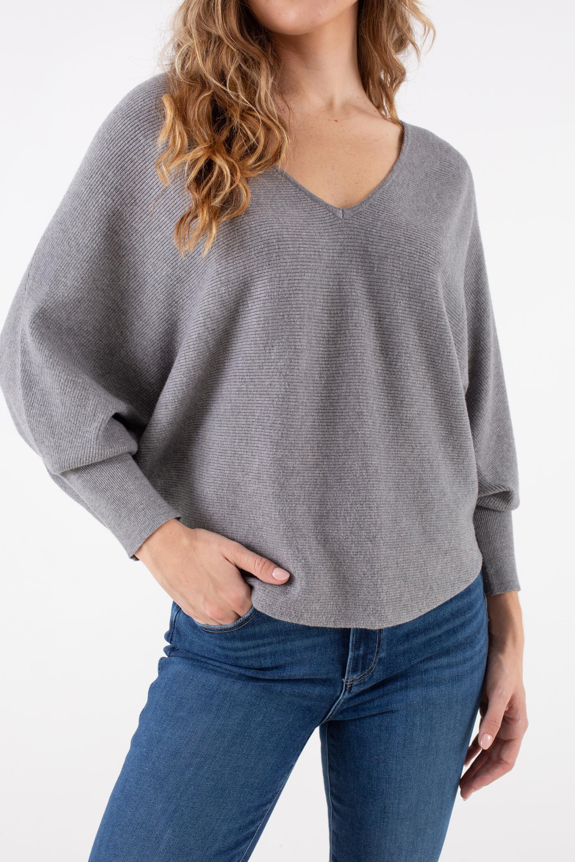 A woman models the Kerisma Dolman Ryu V Neck sweater in Ash Grey—a loose-fitting, long-sleeve V-neck top—paired with blue jeans, standing with one hand in her pocket against a plain white background.