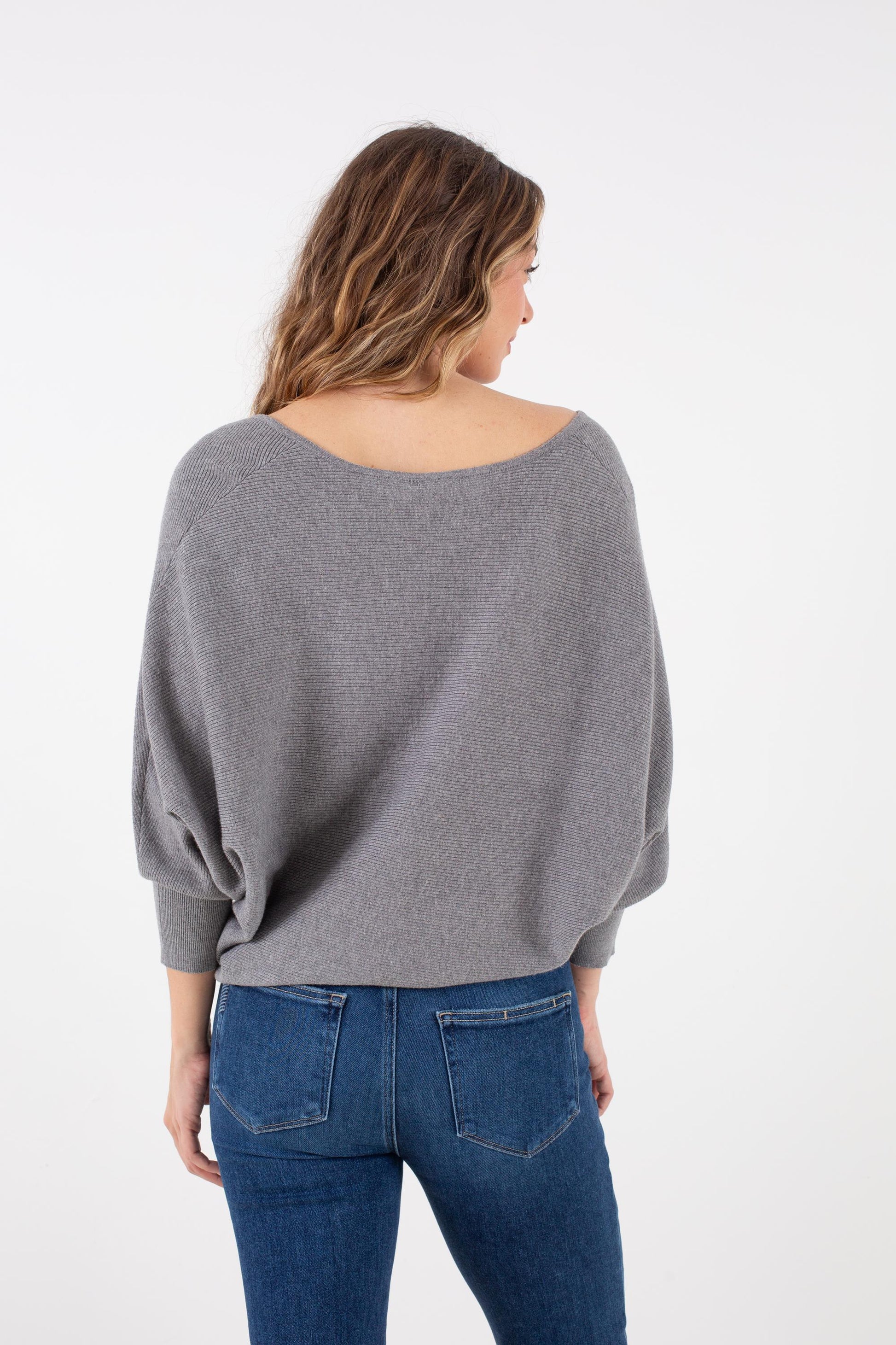 A woman with wavy hair is seen from behind, standing against a plain white background, wearing the Kerisma Dolman Ryu V Neck in Ash Grey paired with blue jeans.