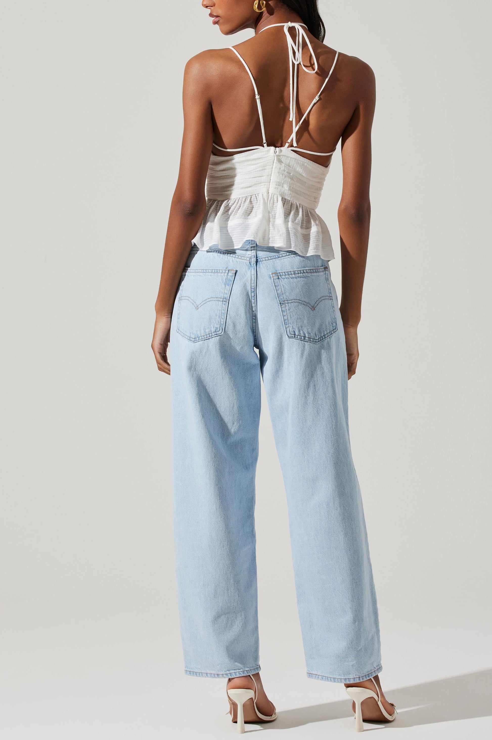 A woman stands with her back to the camera, wearing the ASTR Minka Top—a white strappy design—paired with light blue high-waisted jeans and white heeled sandals against a plain light background, creating a chic, effortless style reminiscent of relaxed rayon dress elegance.