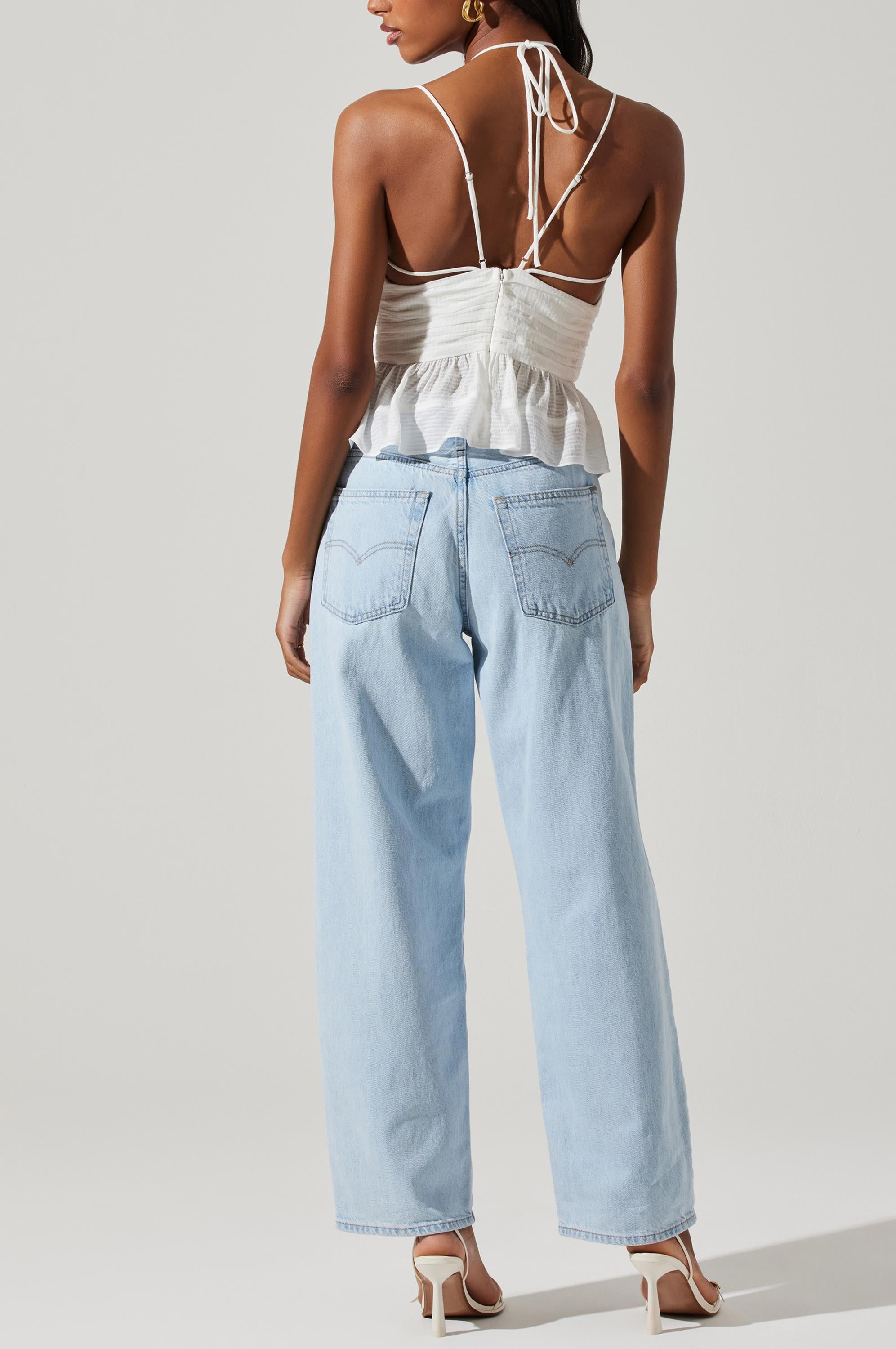A woman stands with her back to the camera, wearing the ASTR Minka Top—a white strappy design—paired with light blue high-waisted jeans and white heeled sandals against a plain light background, creating a chic, effortless style reminiscent of relaxed rayon dress elegance.