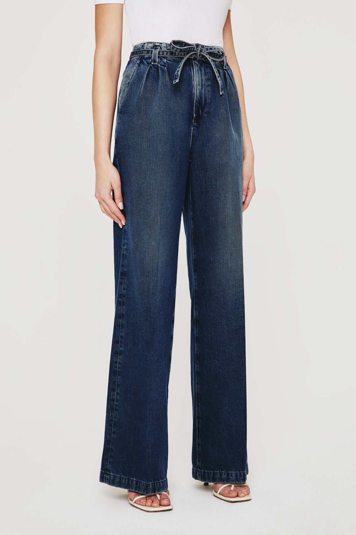 A person wears AG Jeans' Belted Deven In King Street—relaxed fit, high-waisted dark blue jeans with a belted paperbag waist—paired with a white top and open-toe sandals.