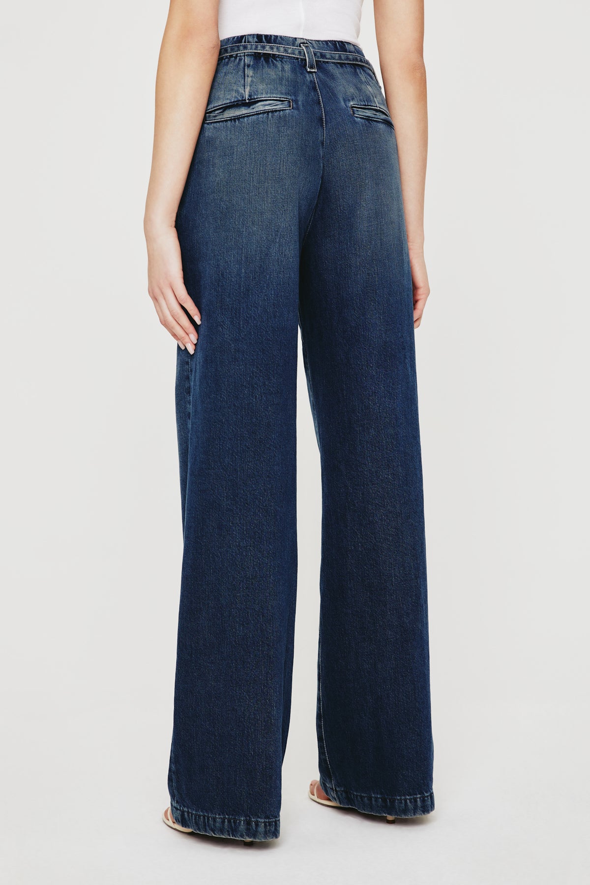 A person is shown from the back wearing AG Jeans' Belted Deven In King Street—high-waisted, relaxed fit, wide-leg blue jeans with welt pockets and a slightly faded wash—paired with a white top against a plain white background.