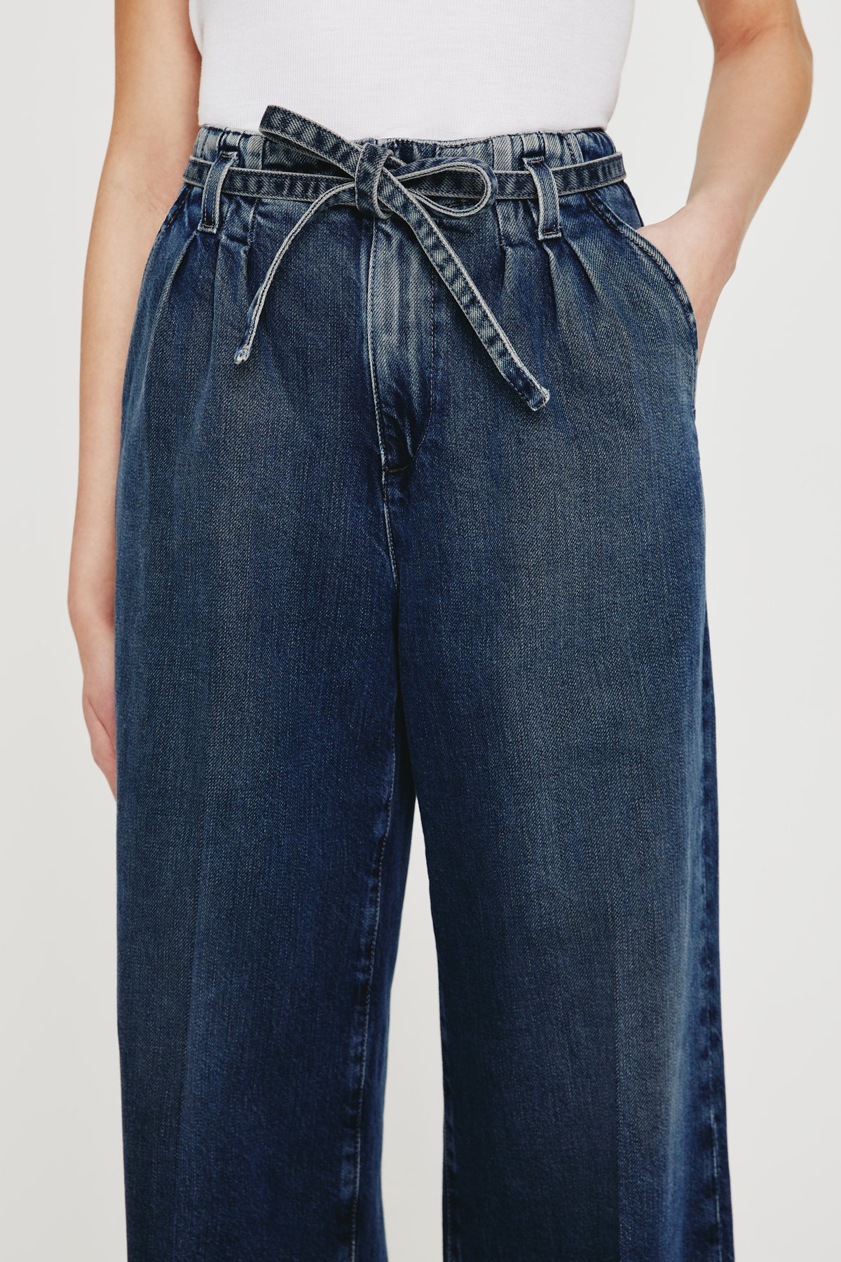 A person wears the AG Jeans Belted Deven In King Street: high-waisted, wide-leg blue jeans with a paperbag waist and matching denim belt tied in a bow. Paired with a white top, the relaxed fit and hands-in-pockets create an effortless style.