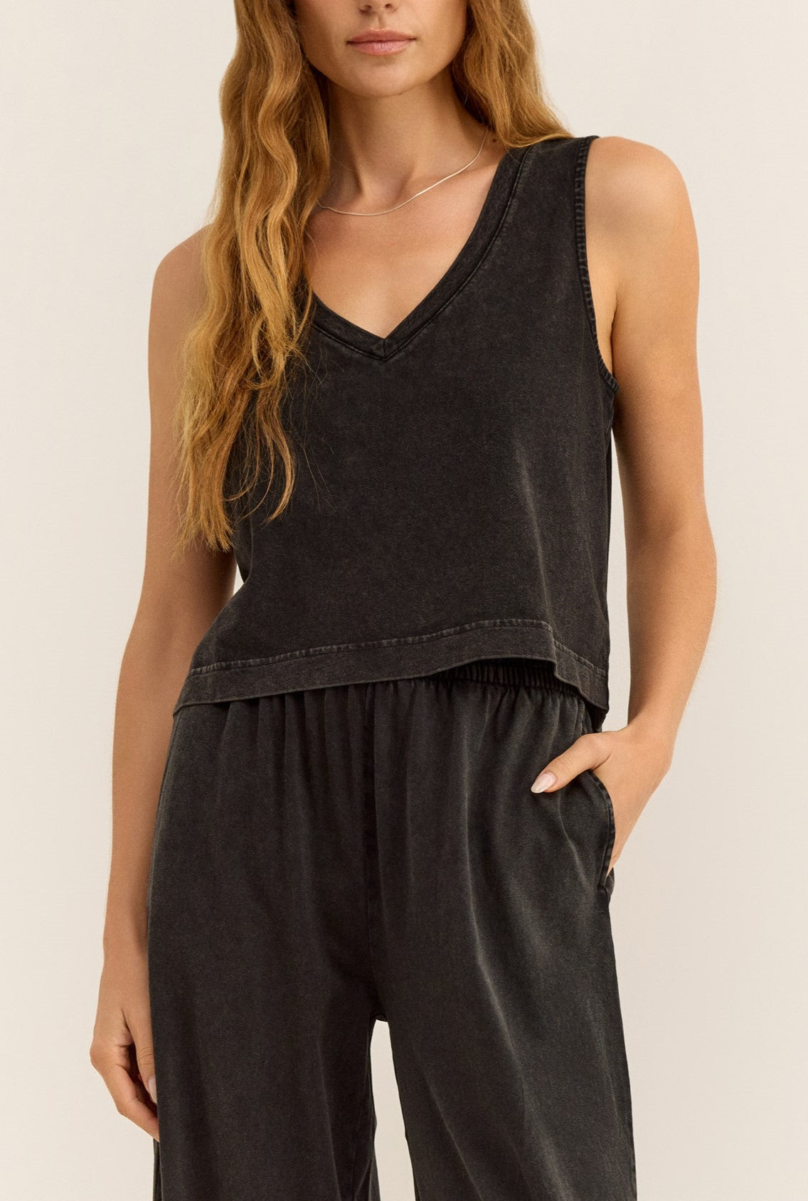 A woman wears the Z Supply Sloane V-neck Top in Black, a sleeveless cotton jersey tank, paired with matching black pants. She stands against a plain light background with one hand in her pocket.