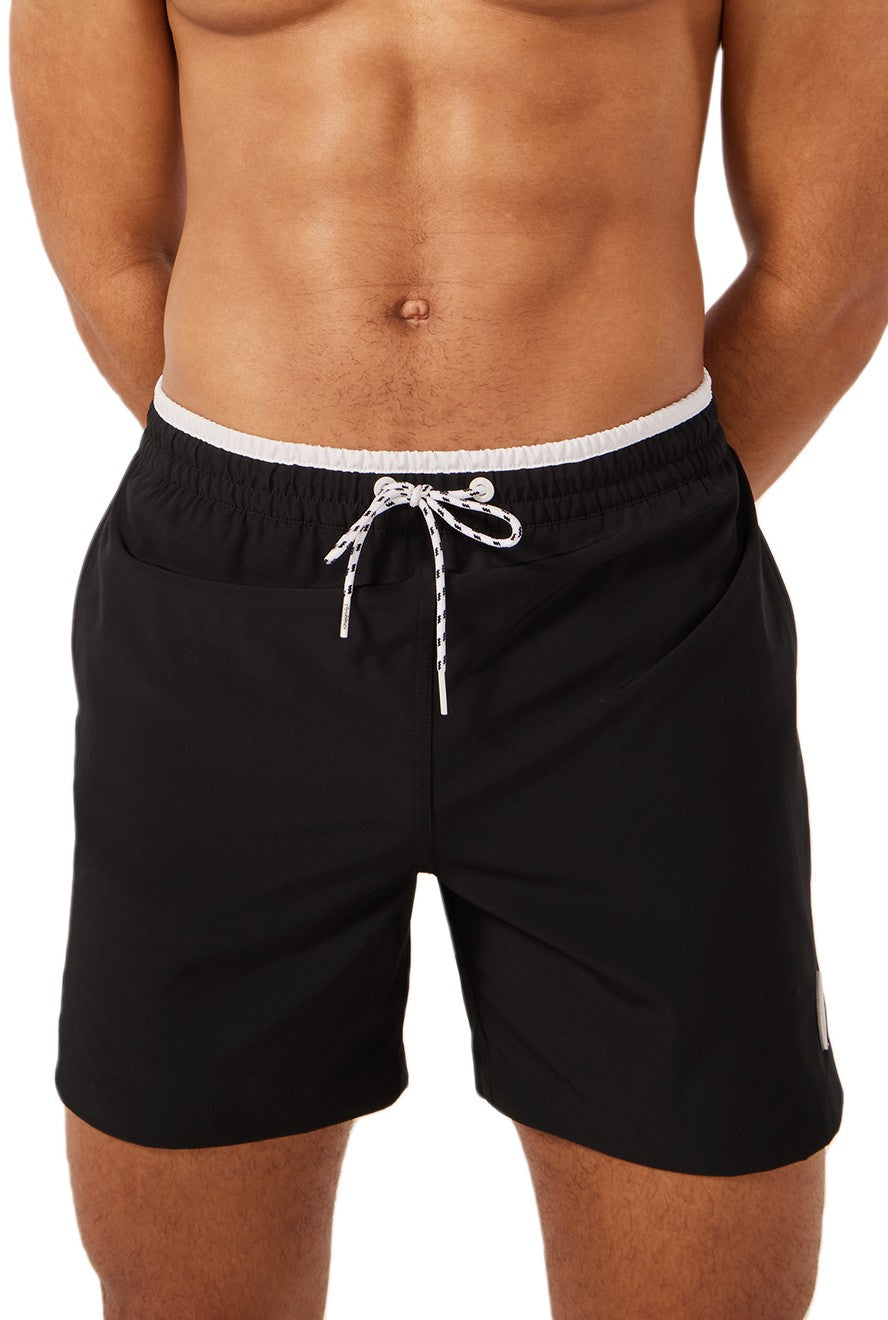 A person stands with hands behind their back wearing The Capes 5.5 Inch Unlined black swim trunks by Chubbies, featuring a white waistband, white drawstring, and quick-dry fabric; the image shows their torso and upper legs.