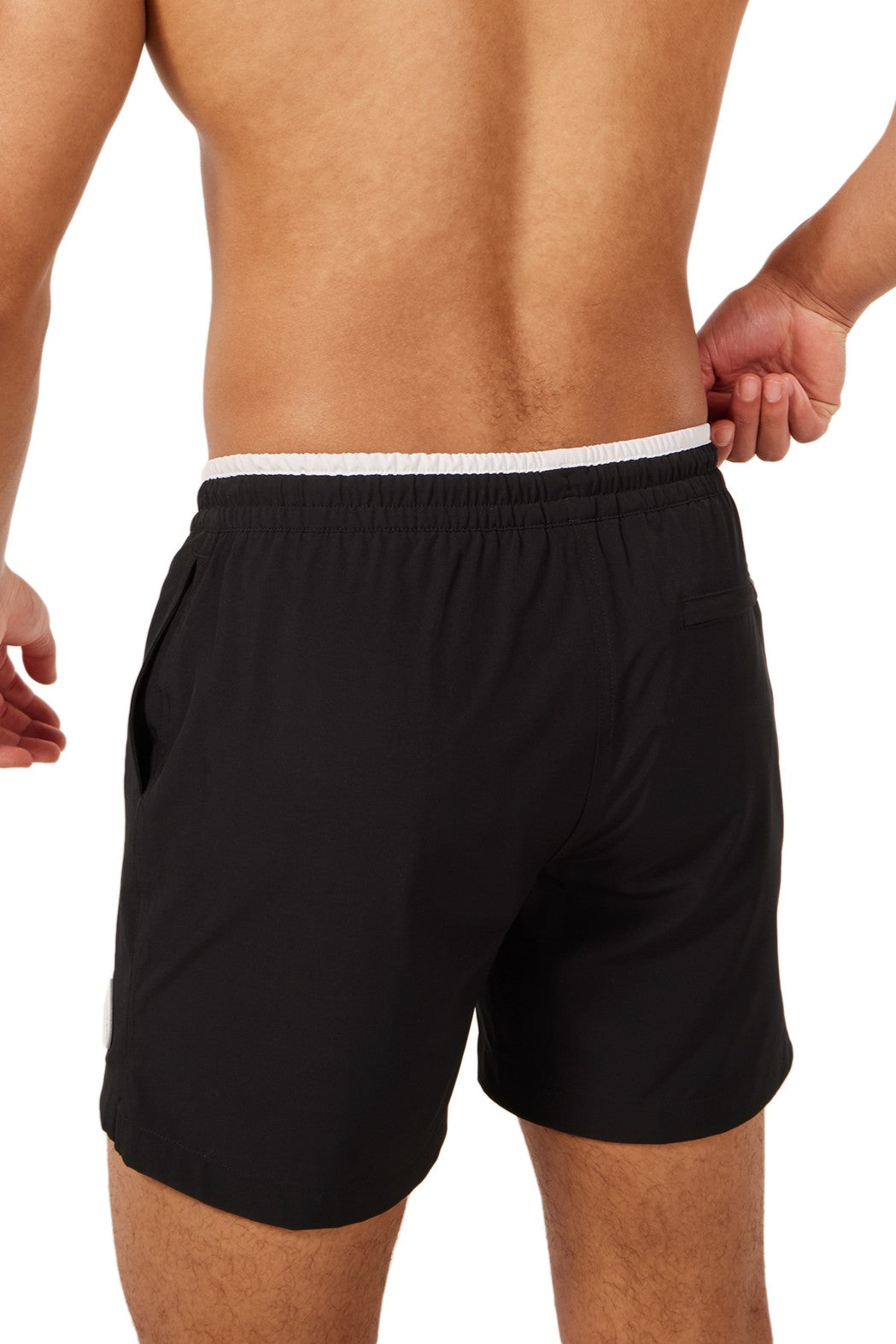 A shirtless person, shown from the back against a white background, adjusts the waistband of black Chubbies The Capes 5.5 Inch Unlined athletic shorts featuring a white waistband and quick-dry fabric.
