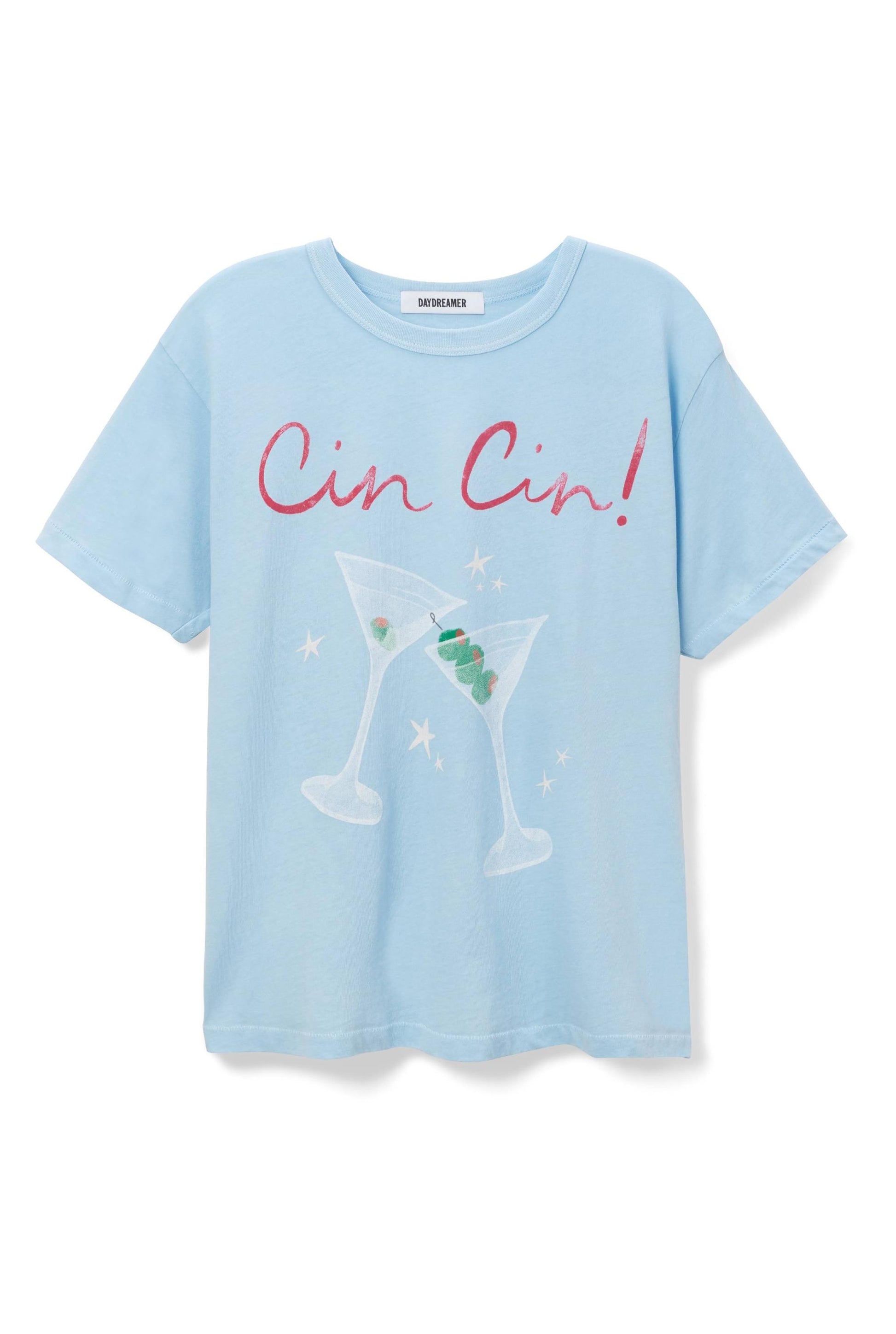 The Cin Cin Ringer Tee by Daydreamer LA is a light blue shirt with two illustrated martini glasses clinking, green olives inside, stars around them, and "Cin Cin!" in large pink script above.