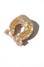 The Solar Eclipse Handpainted Horseshoe Claw Clip features a pearly pink finish with gold glittery sections, blue dots, and star stickers, displayed against a white background.
