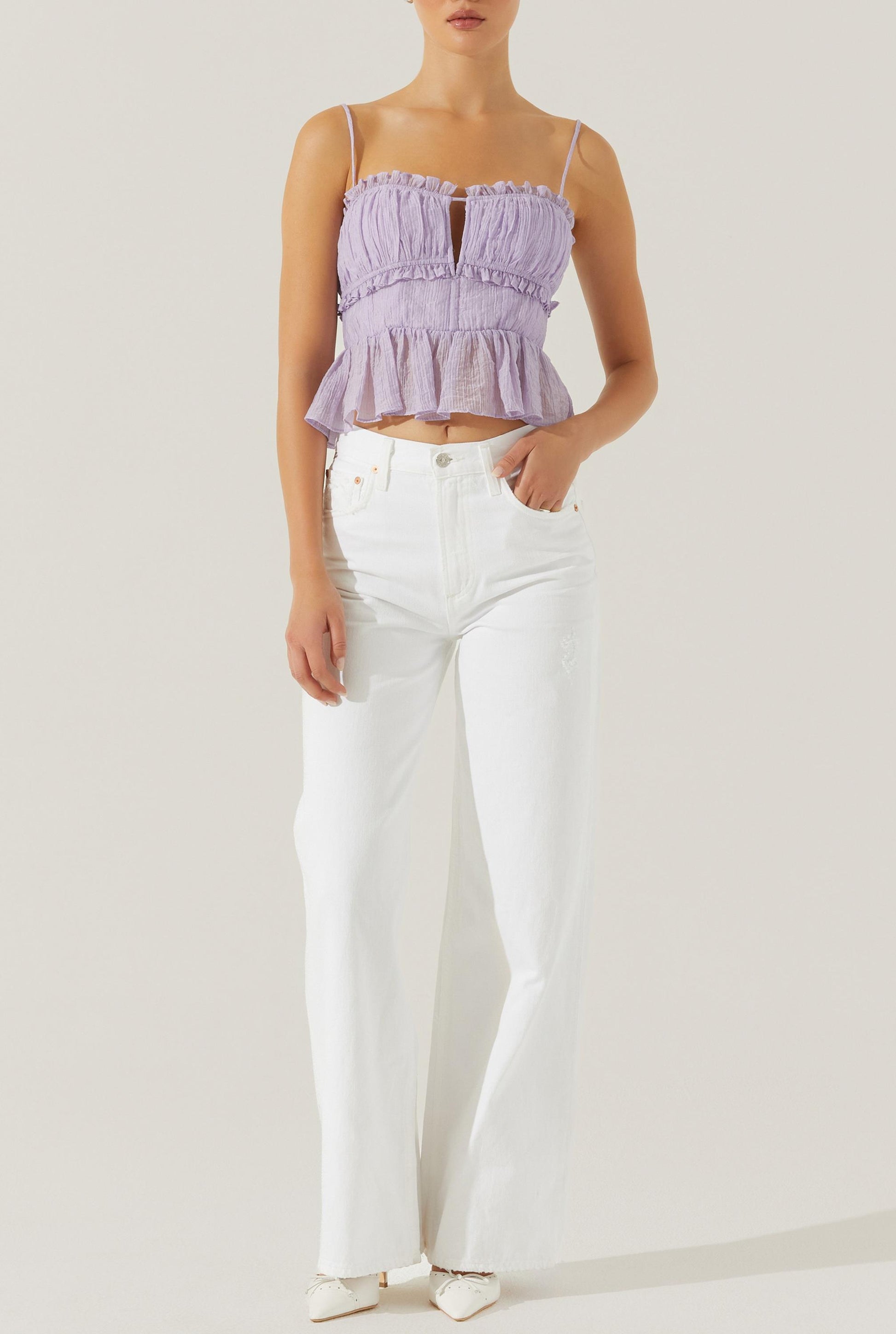 A woman wearing the Sylvan Top by ASTR—a sleeveless light purple crepe peplum top featuring adjustable spaghetti straps and a milkmaid bust—paired with high-waisted white wide-leg jeans and white pointed shoes, stands against a plain light background.