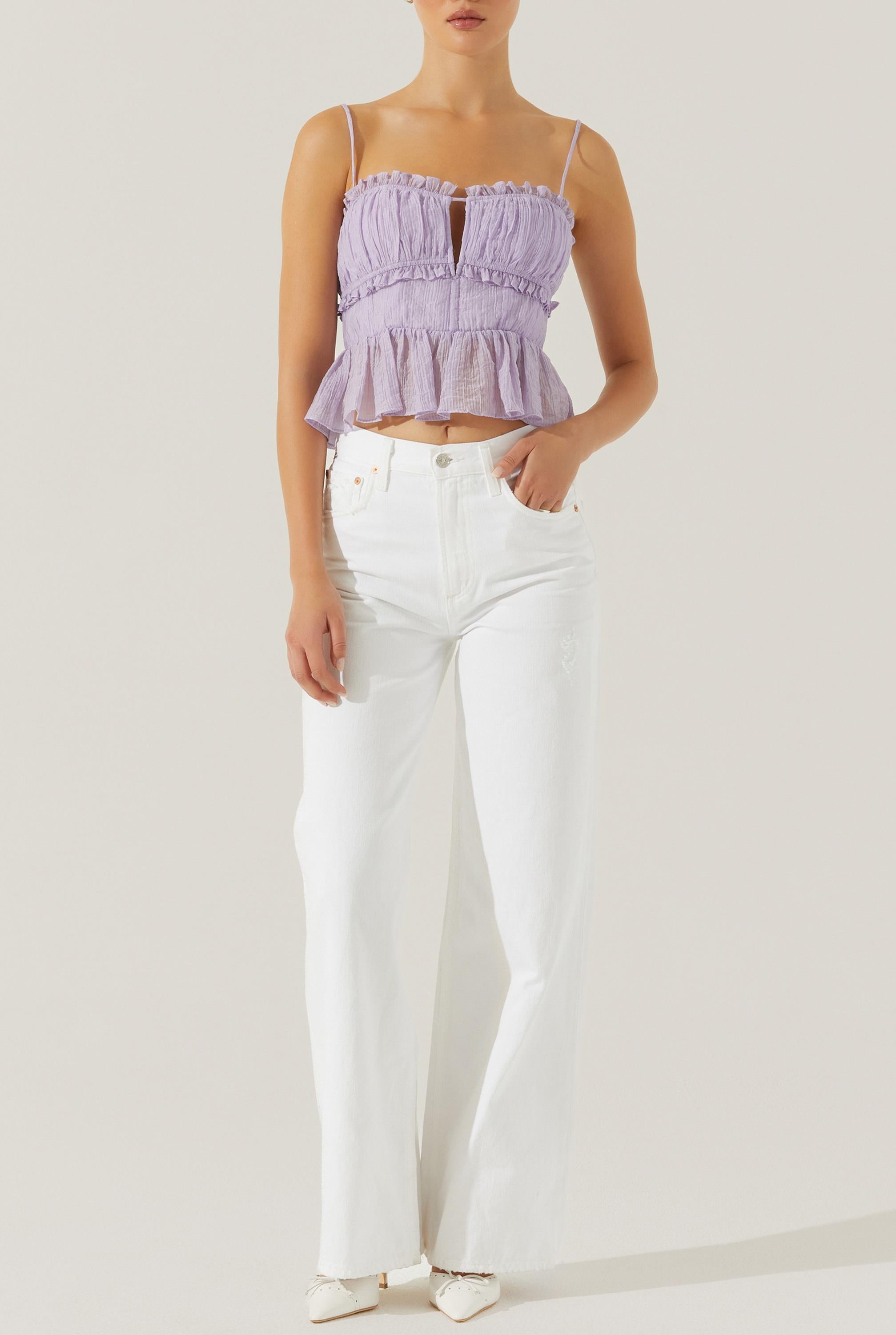 A woman wearing the Sylvan Top by ASTR—a sleeveless light purple crepe peplum top featuring adjustable spaghetti straps and a milkmaid bust—paired with high-waisted white wide-leg jeans and white pointed shoes, stands against a plain light background.