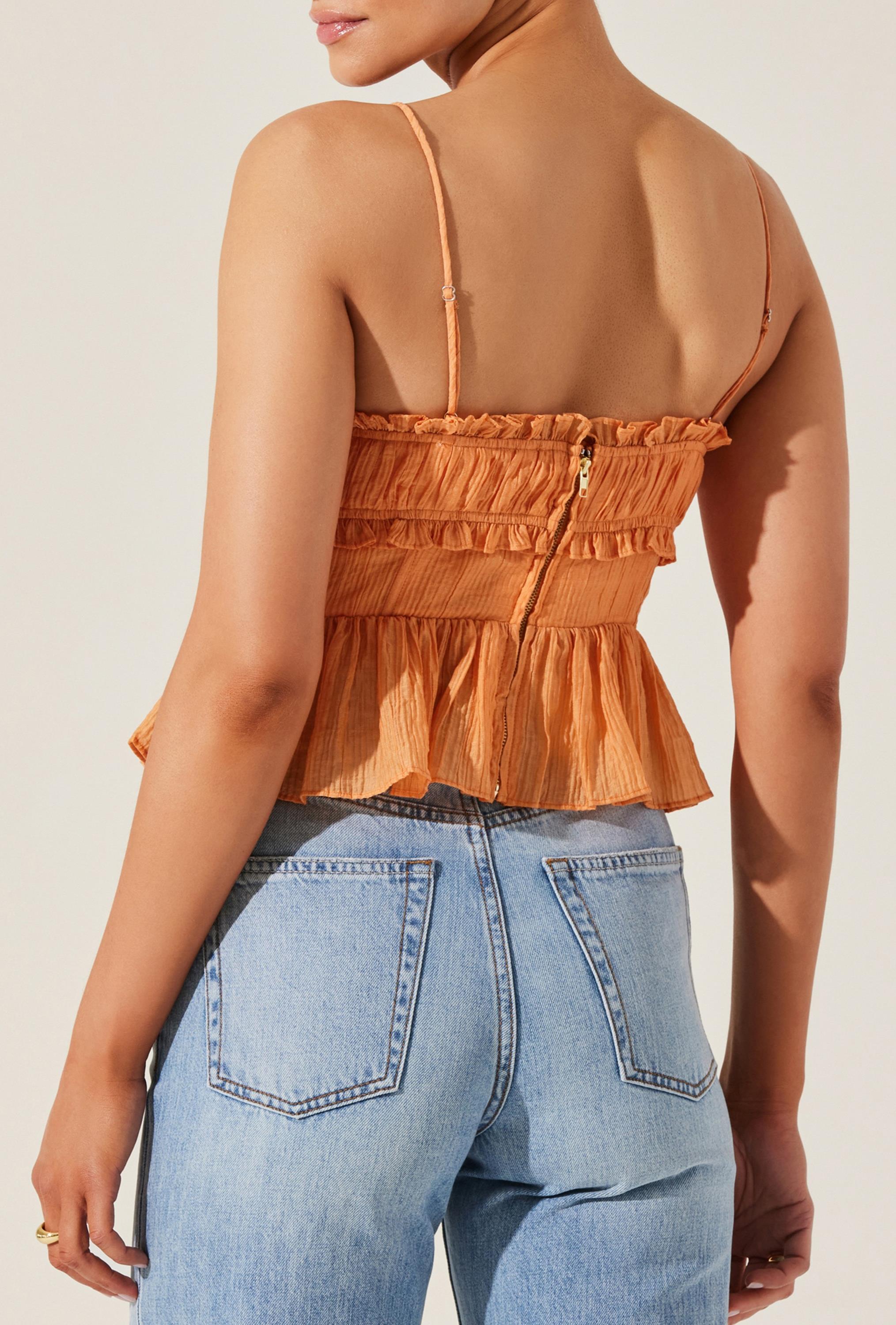 A woman is wearing the Sylvan Top by ASTR, a sleeveless orange crepe peplum style featuring ruffle trim and adjustable spaghetti straps, paired with light blue high-waisted jeans. The image is taken from behind, showcasing the top's zipper detail and the jeans' back pockets.