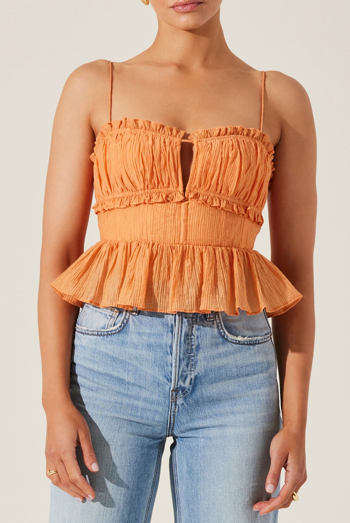 A person wearing the ASTR Sylvan Top—a sleeveless orange crepe peplum top with thin straps and ruffle trim—paired with light blue high-waisted jeans.
