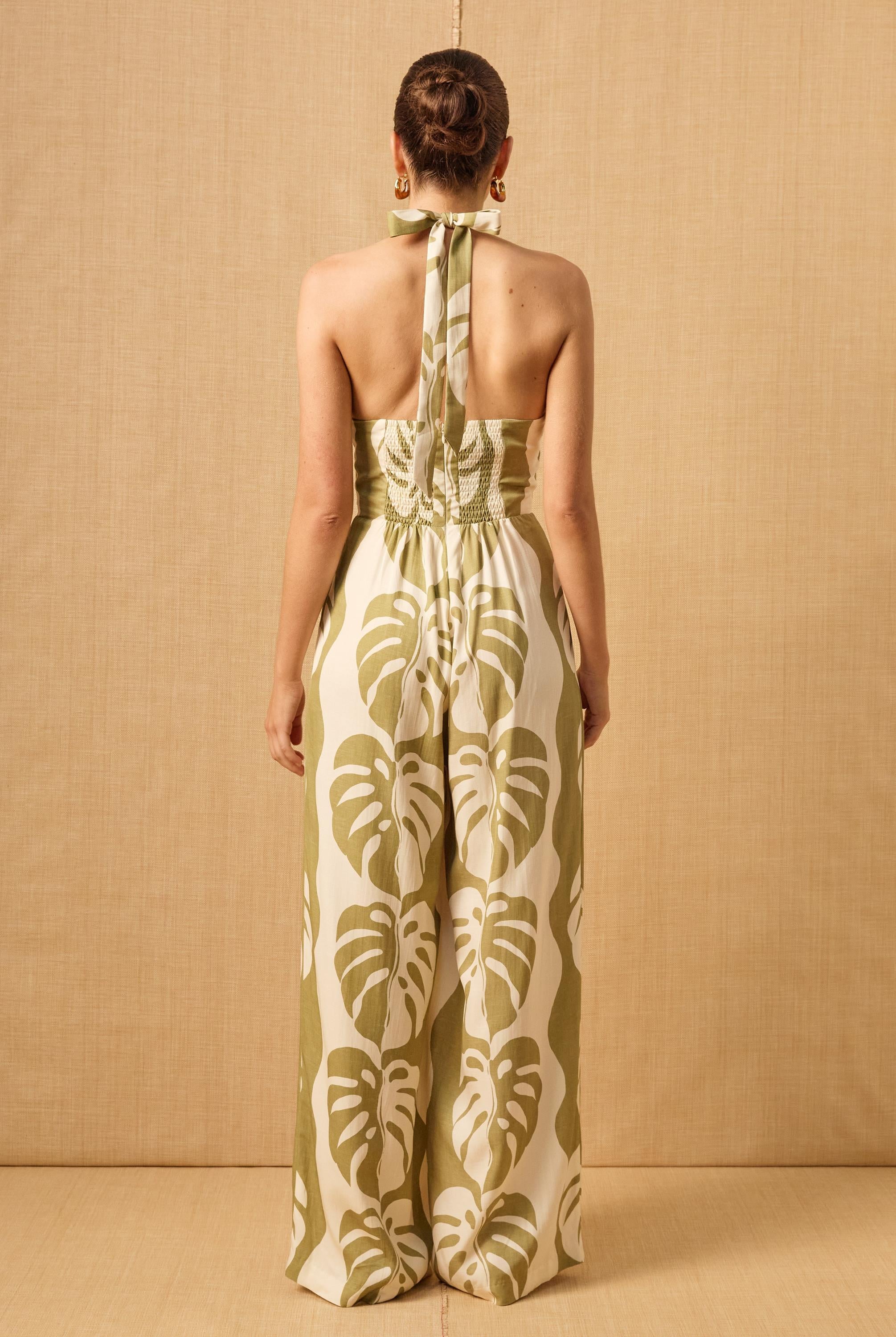 A woman with a neat bun stands with her back to the camera wearing the Mon Renn Midsummer Palm Jumpsuit, a blended linen halter-neck style with green and white monstera print and wide legs, against a beige textured background.