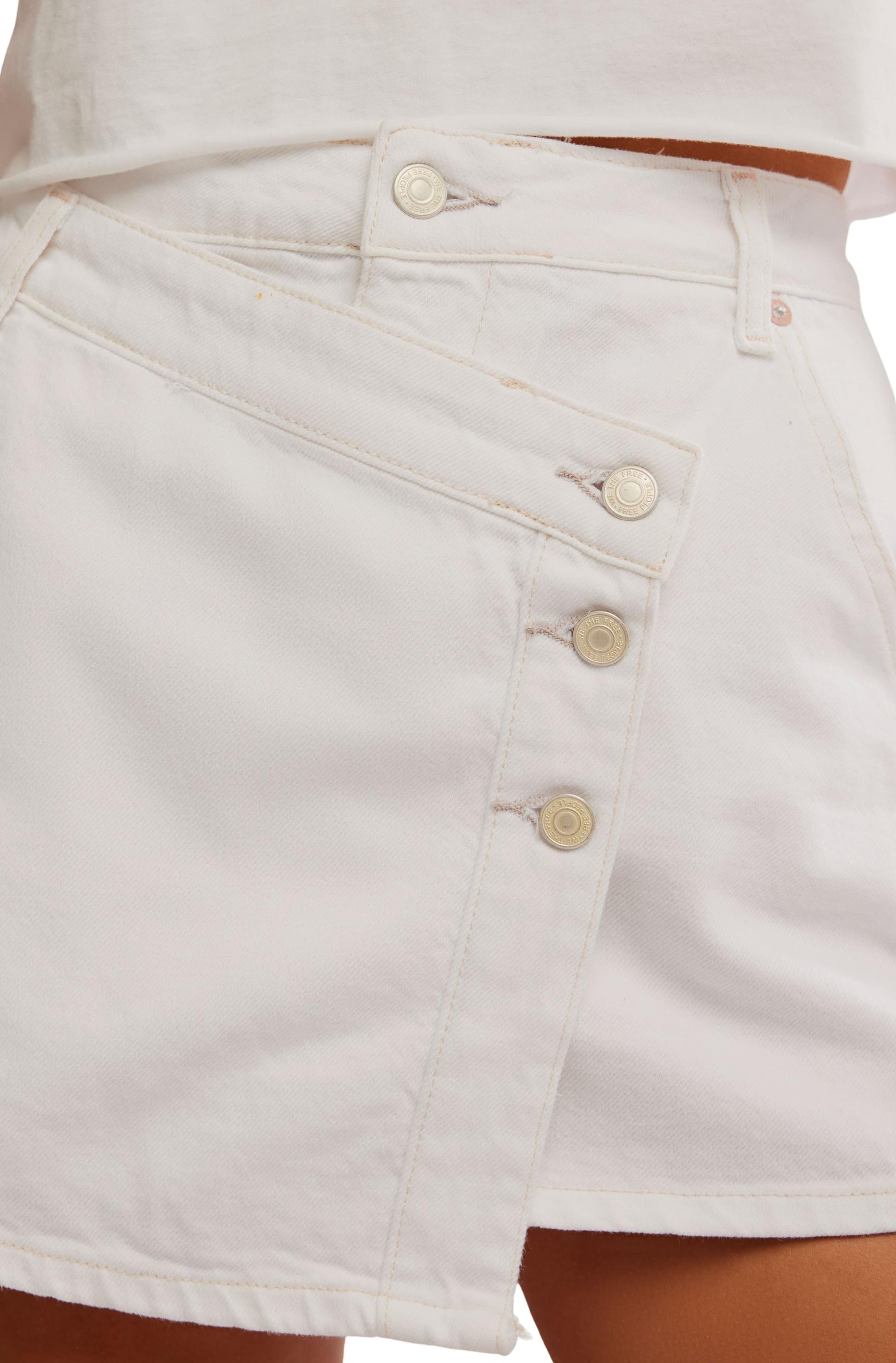 Close-up of a person wearing the Wynne Denim Skirt by Free People, featuring a white wash, asymmetrical front panel, and multiple buttons. The skirt is paired with a partially visible white top.