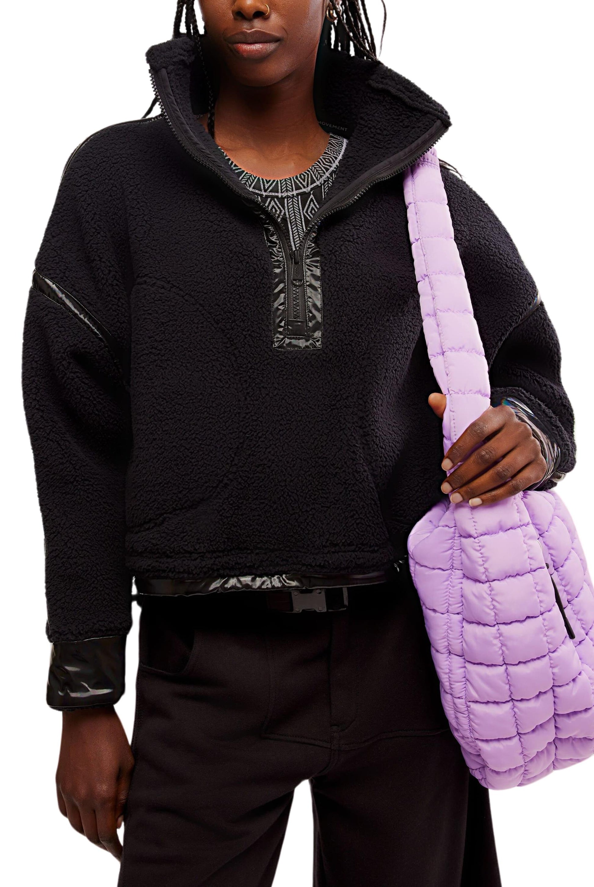 A person wears FP Movement’s Mountain High Shine 1/2 Zip pullover—black and textured with shiny trim, a high collar, and zip details—with dark pants, holding a large quilted lavender shoulder bag.