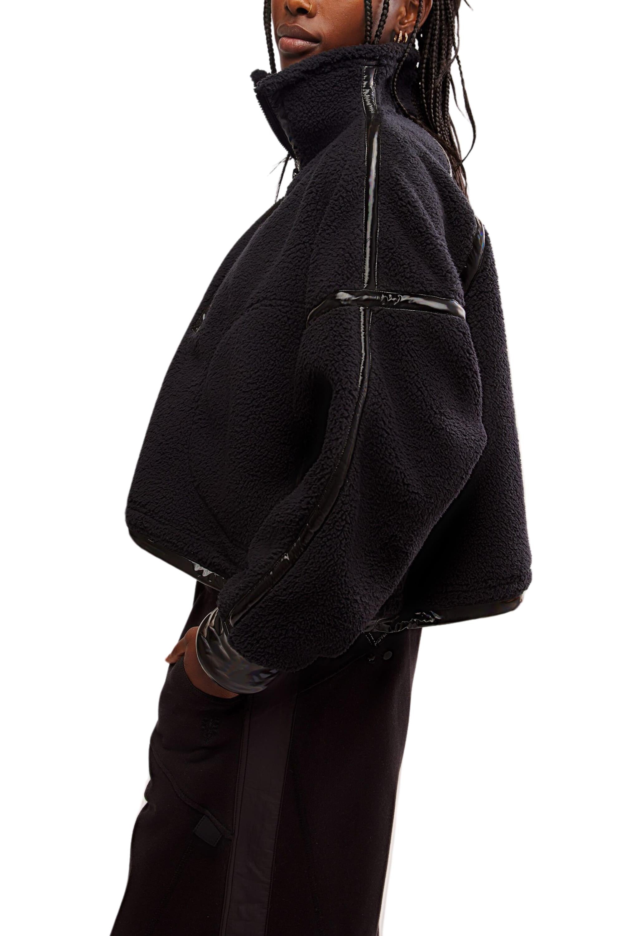 A person wears the FP Movement Mountain High Shine 1/2 Zip, a black oversized sherpa jacket with shiny trim and a high collar, standing sideways with hands in pockets for a stylish, modern look.