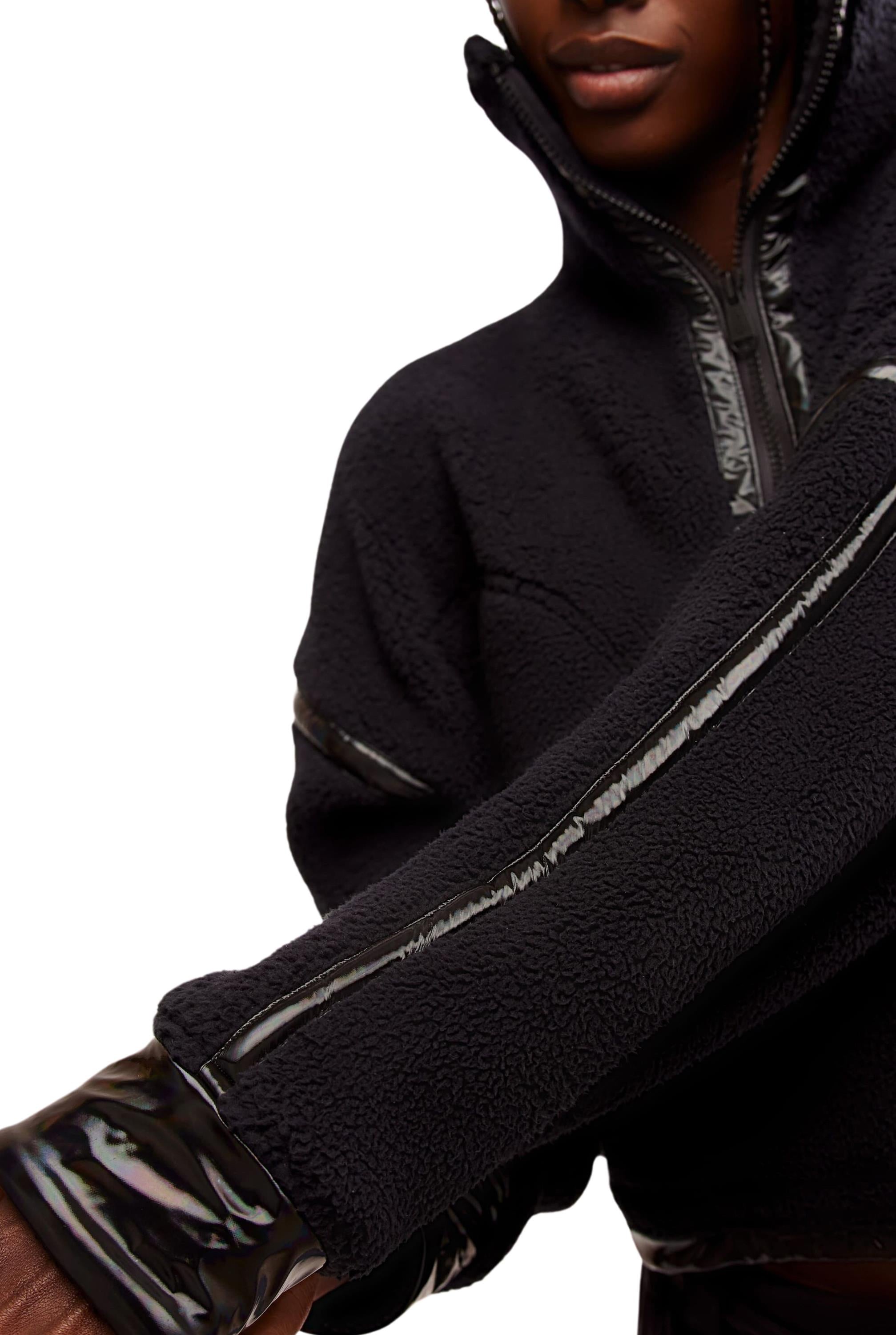 A person wears the FP Movement Mountain High Shine 1/2 Zip, a black textured jacket with shiny, vinyl-like trim on the sleeves and zipper, shown from shoulders to waist against a white background.