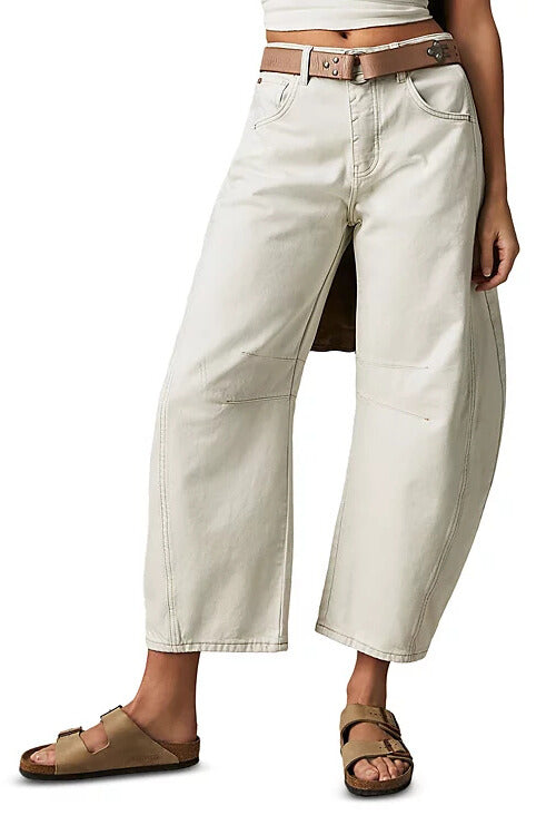 Someone models the Free People Good Luck Mid Rise Barrel jeans in cream—mid-rise, barrel-leg style, cropped at the ankle—paired with a brown belt and tan sandals, one leg forward, set against a white background.