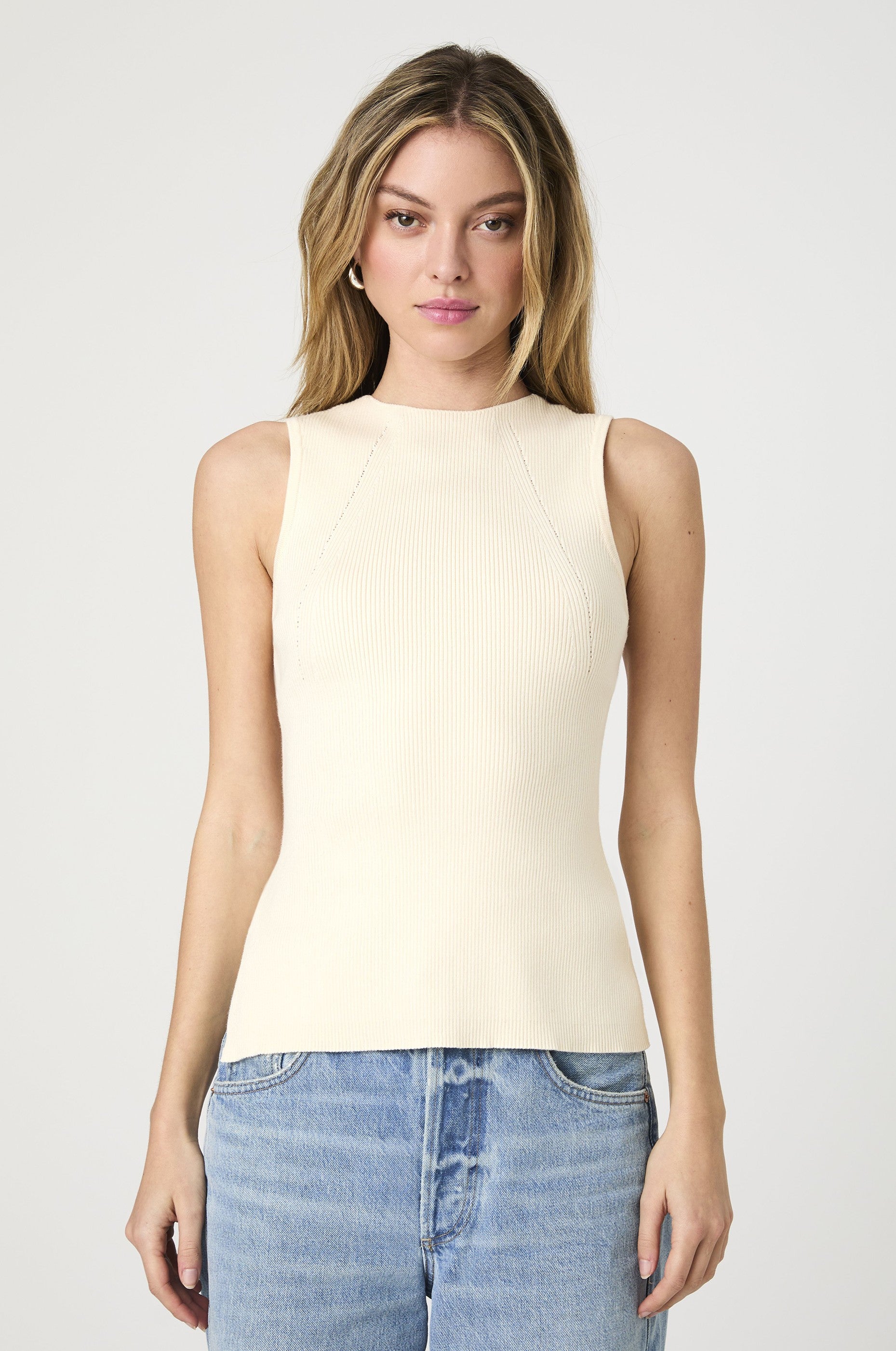 A woman with light brown hair wears the French Connection Mozza Rib Tank Top in Ecru, paired with light blue jeans, and stands against a plain, light background.