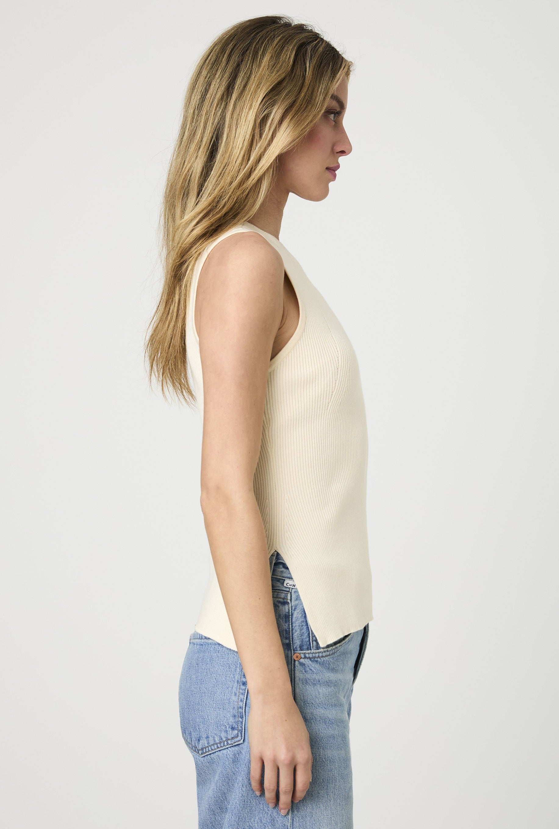 A person with long blonde hair stands in profile against a plain white background, wearing the French Connection Mozza Rib Tank Top in beige and light blue jeans.