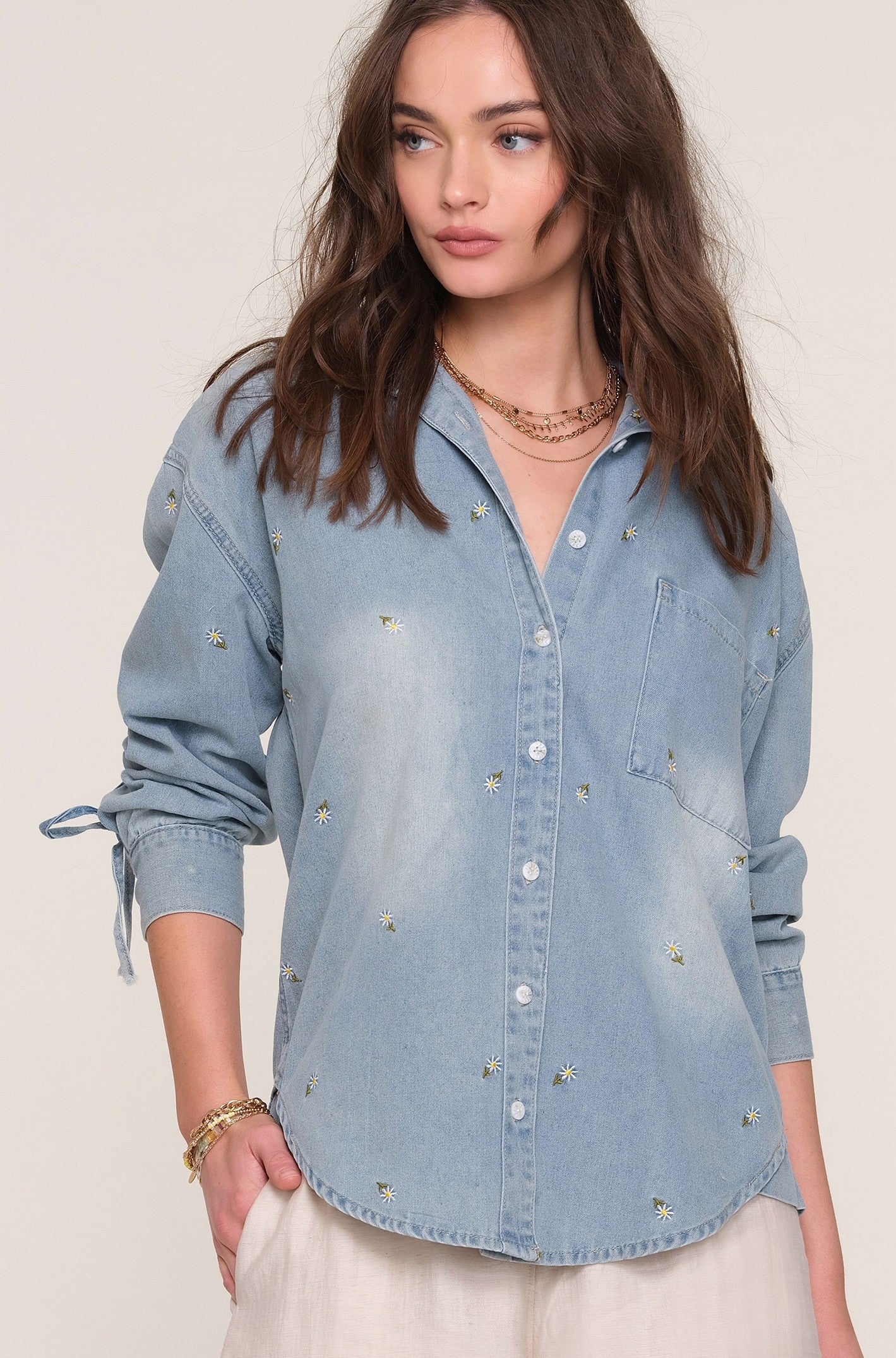 A woman with long brown hair wears the Heartloom Harper Shirt, a light blue denim top with relaxed fit, button-down closure, and small embroidered flowers, styled with white pants and layered jewelry against a plain background.