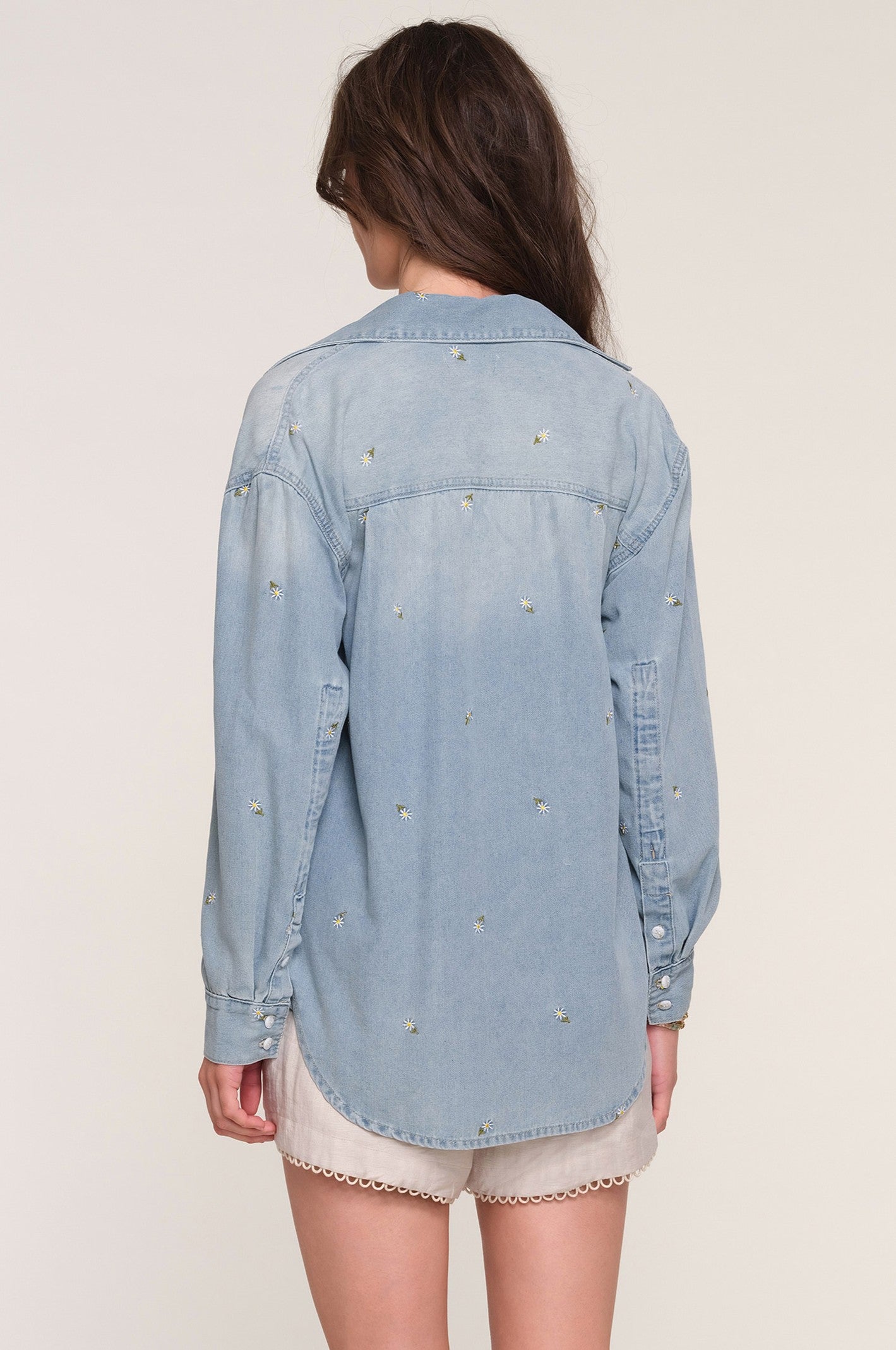 A woman with long brown hair, seen from behind, wears Heartloom's Harper Shirt—a relaxed fit light blue denim top with embroidered details and button-down closure—paired with light shorts that have scalloped edges. The background is plain off-white.