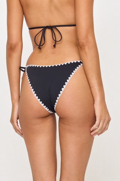 A person wearing the LSpace Black/Cream Zahra Bottom, a black mid-rise bikini with cream zigzag trim and side ties, is shown from the back with one arm relaxed by their side. The bottom offers classic coverage.