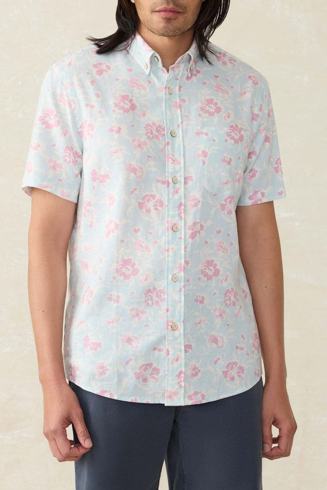 A person wears the Faherty Short Sleeve Breeze Shirt in light blue with a pink floral pattern and dark pants, standing against a light background. Their face is partially visible.