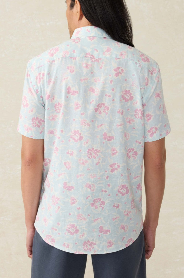 A person with long dark hair, seen from behind, wears the Faherty Short Sleeve Breeze Shirt in light blue with a pink floral pattern and dark pants. The background is a neutral light beige color.