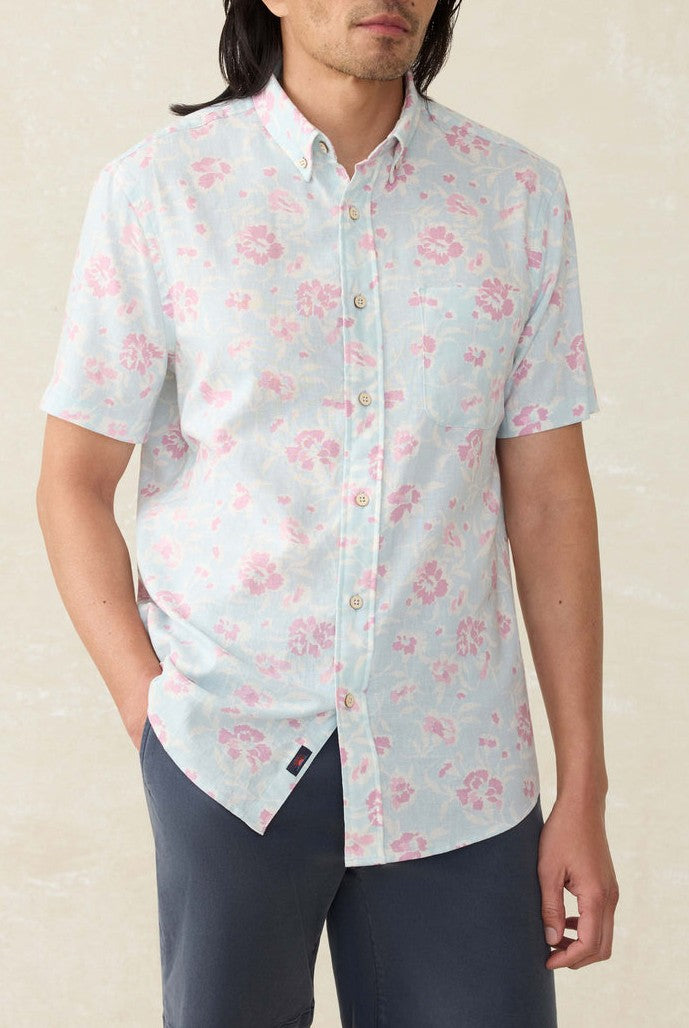 A man stands against a neutral background wearing Faherty's Short Sleeve Breeze Shirt in light blue, featuring a relaxed fit and pink floral pattern, paired with dark blue pants and one hand in his pocket.