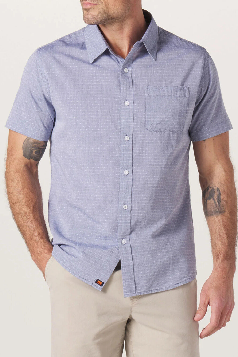 A man stands against a plain background wearing The Normal Brand’s Freshwater Button Up Shirt in BLUE DOBBY—a light blue, short-sleeve button-up crafted from 100% cotton with flat felled seams, a classic button placket, and a chest pocket—paired with beige pants. Tattoos are visible on both arms.