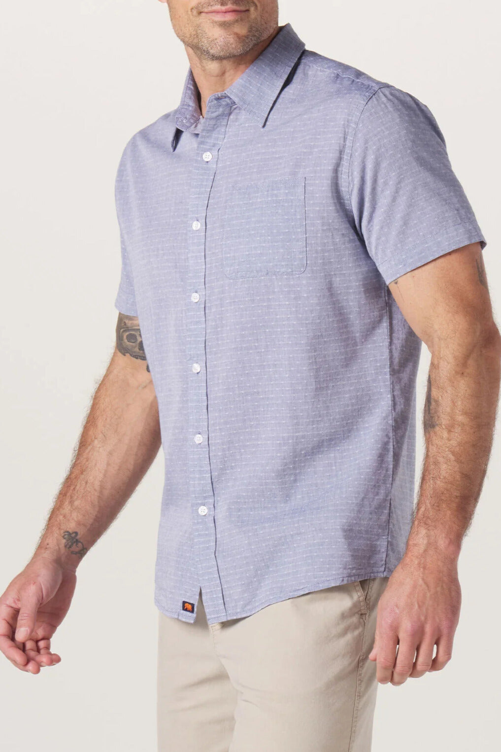 A man wearing The Normal Brand Freshwater Button Up Shirt—a light blue short-sleeve shirt featuring a chest pocket, button placket, and shirttail hem—paired with beige pants, stands against a plain light background.