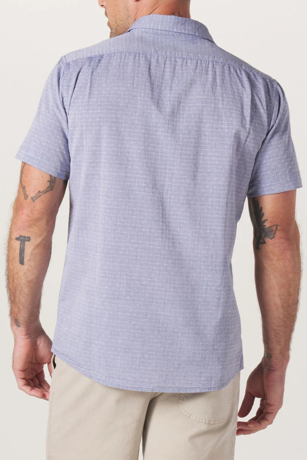A man is shown from the back wearing The Normal Brand's Freshwater Button Up Shirt in light blue, a short-sleeve 100% cotton shirt with a classic button placket, paired with beige shorts. His tattoos are visible on both arms, and the image is set against a plain white background.