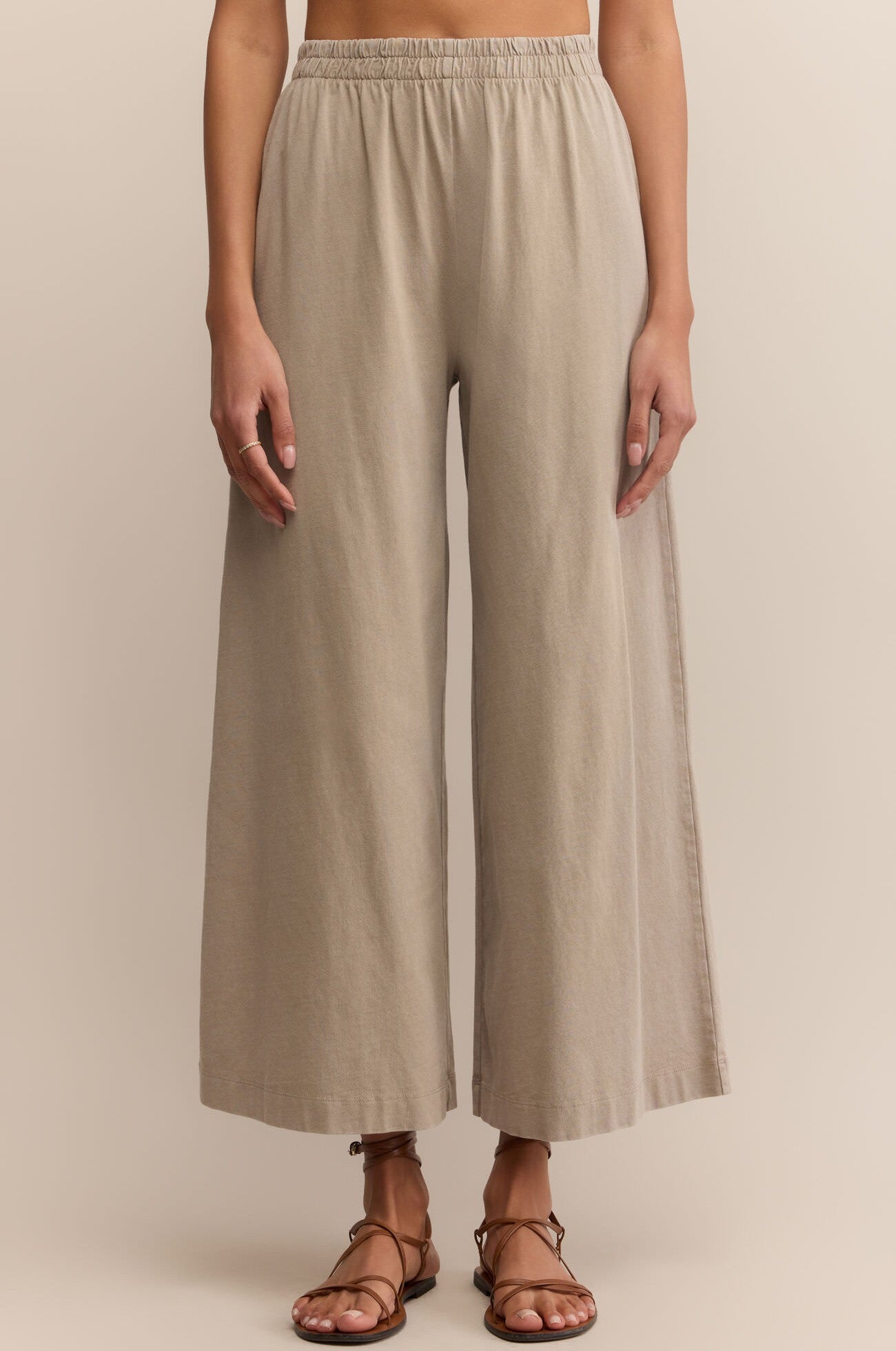 Wearing Z Supply’s Scout Jersey Crop Flare Pant in beige with an elastic waistband and brown strappy sandals, a person stands against a neutral background, their upper body mostly out of frame.