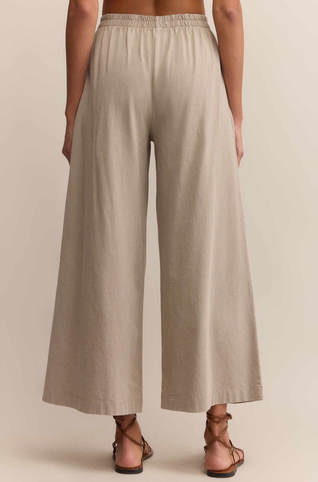A person shown from the back, wearing Z Supply's Scout Jersey Crop Flare Pant in light beige cotton with a relaxed fit and wide flare silhouette, paired with brown strappy sandals against a neutral background.