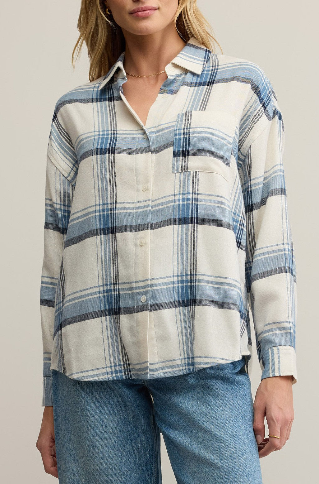 A woman wears the Z Supply River Plaid Button Up in Vintage Indigo—a loose-fitting, blue and white plaid flannel shirt with a collar, long sleeves, button front, and chest pocket—paired with light blue jeans. Her face is partially out of frame.